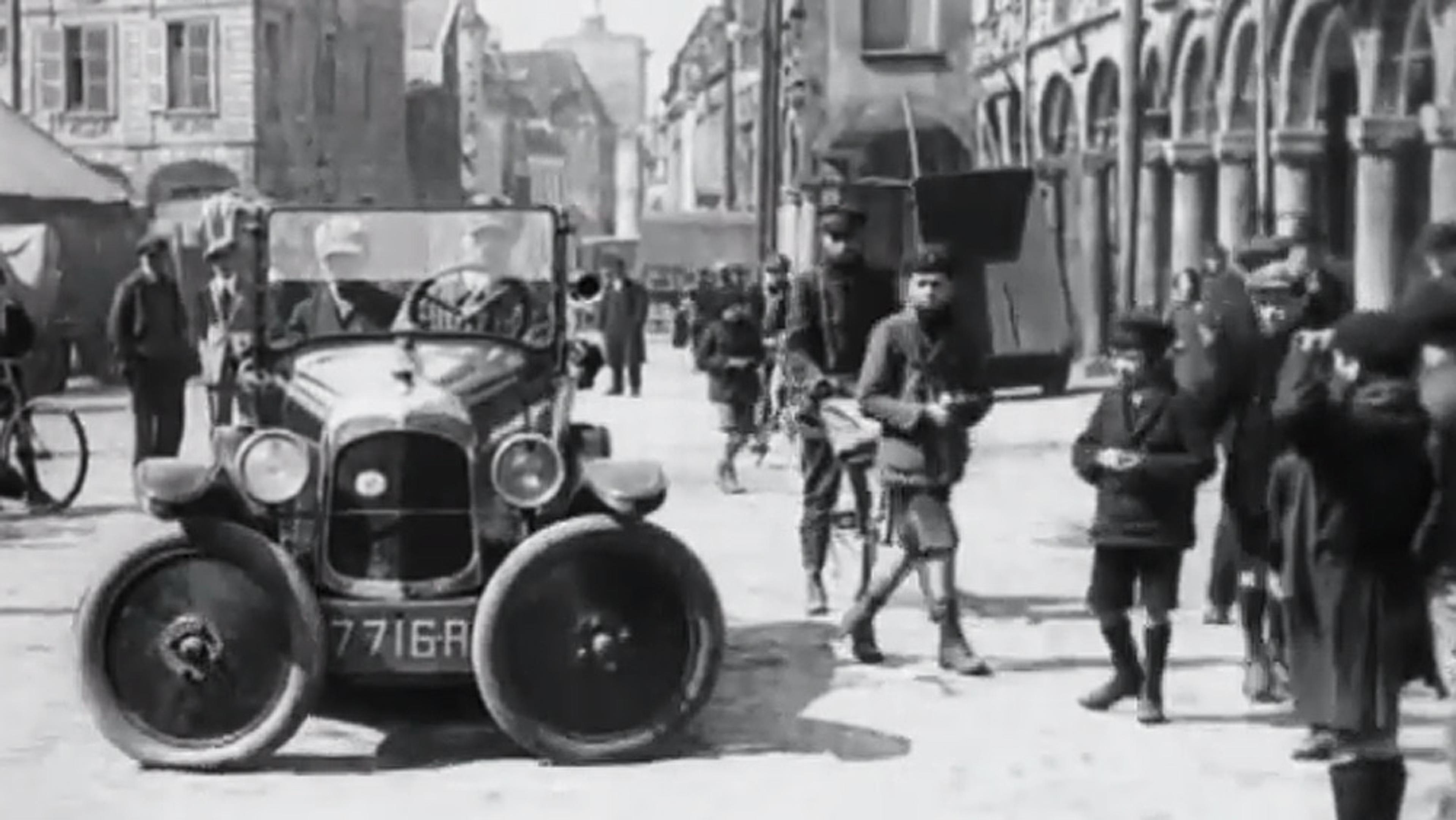 Black and white photo of a vintage car on a bustling city street with pedestrians, including children, and historic buildings in the background.