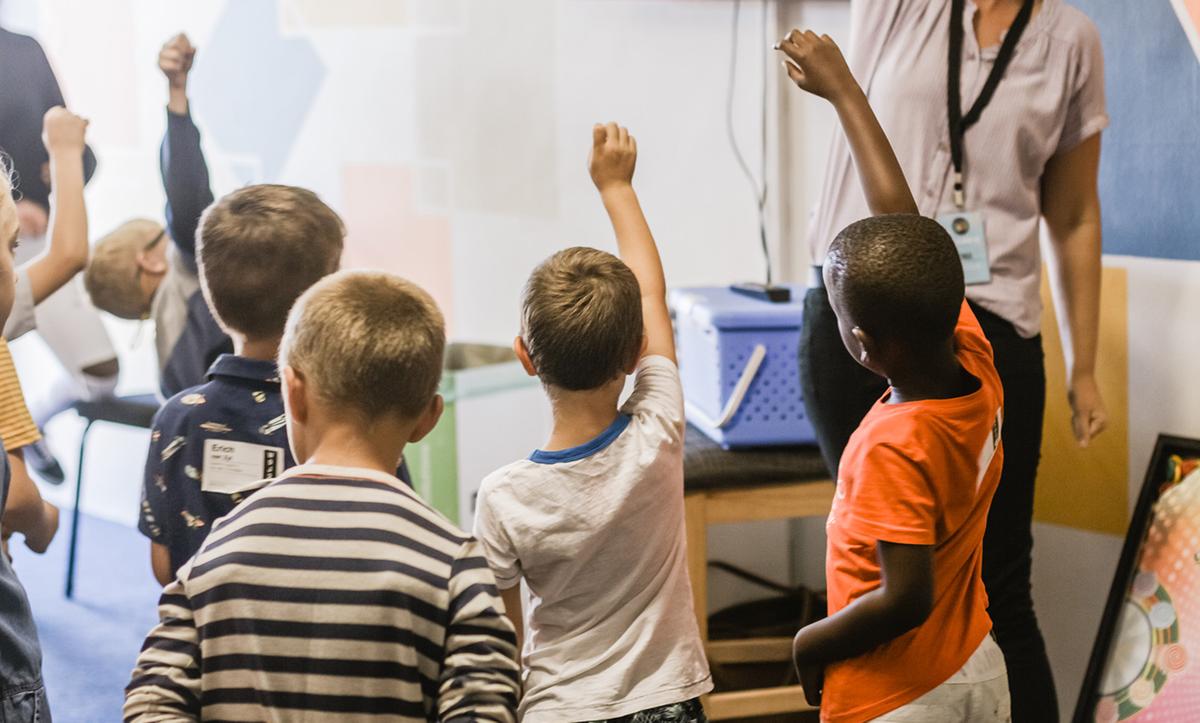 Children in a classroom raising their hands with a teacher at the front. The setting seems to have a relaxed atmosphere.