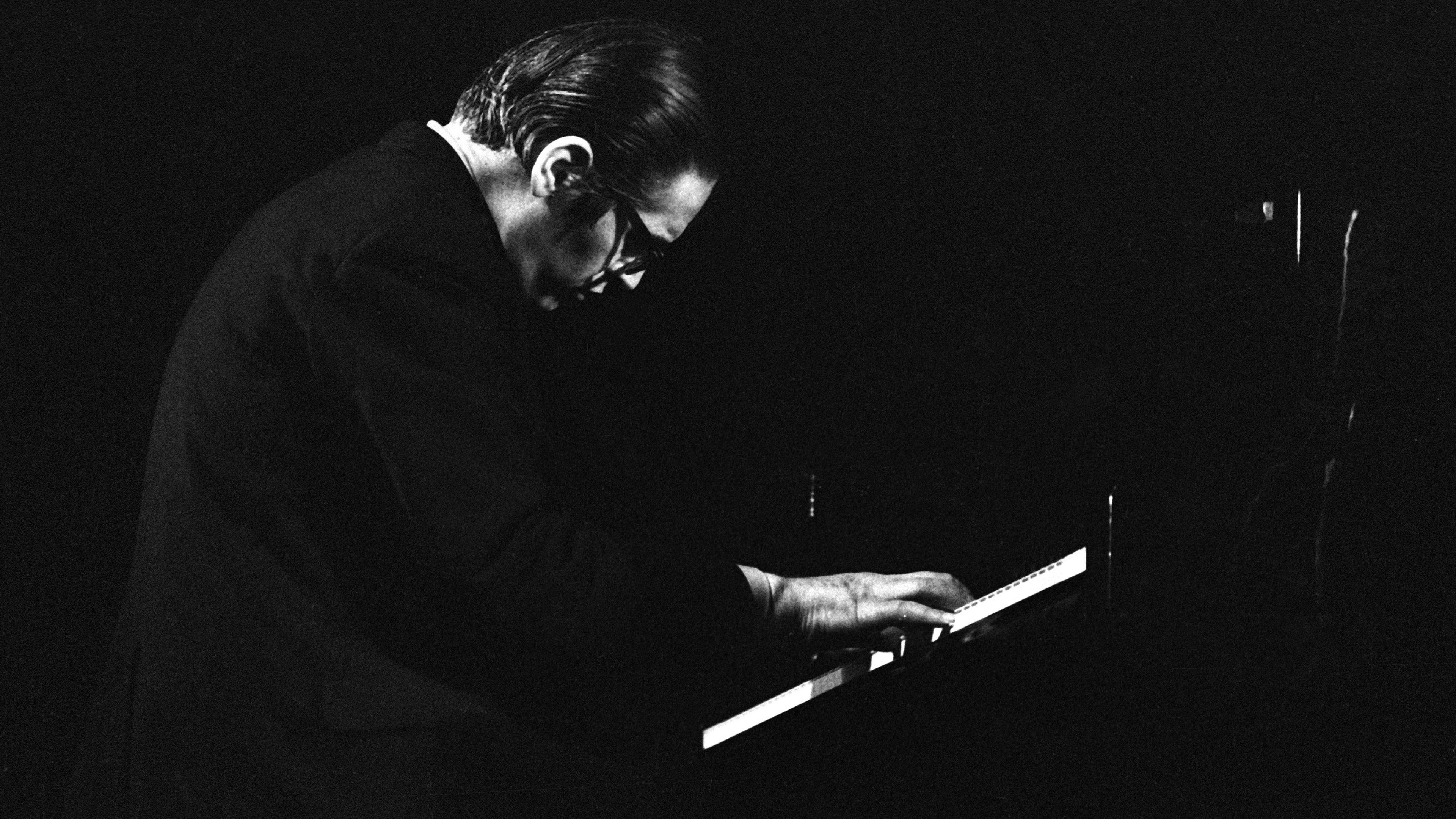 Black and white photo of a man playing piano, with dark background and dramatic lighting.