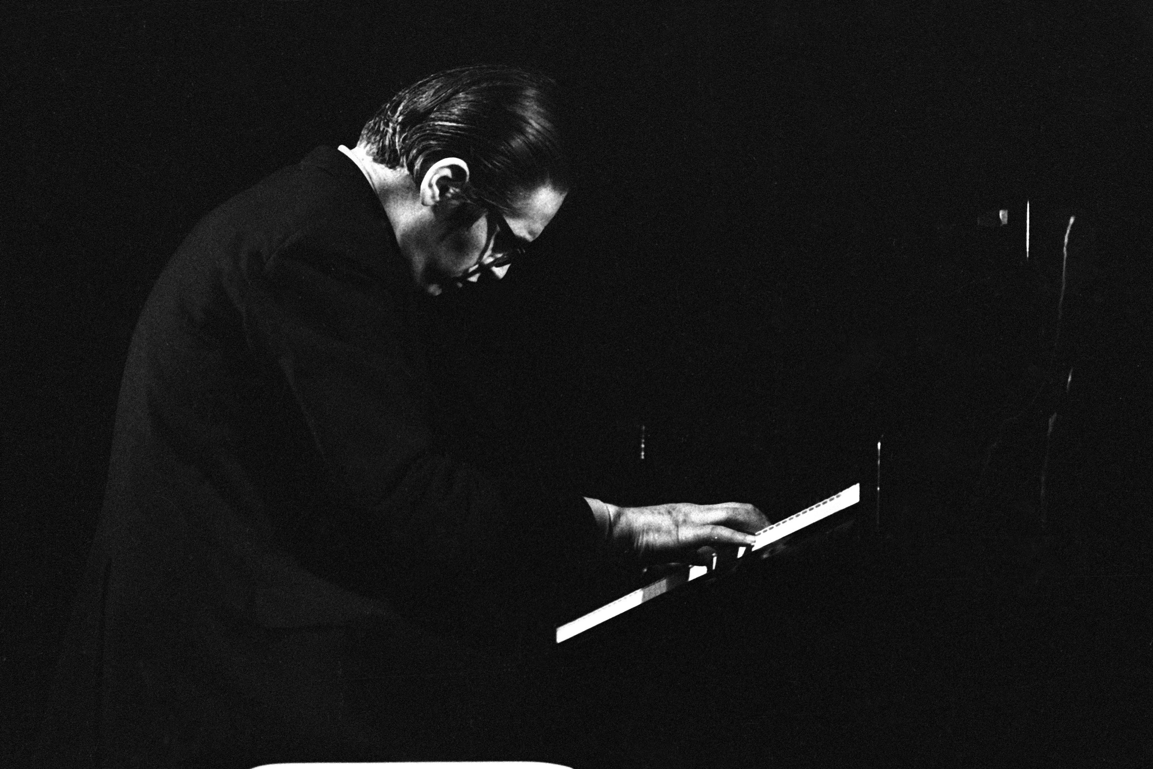 Black and white photo of a man playing piano, with dark background and dramatic lighting.