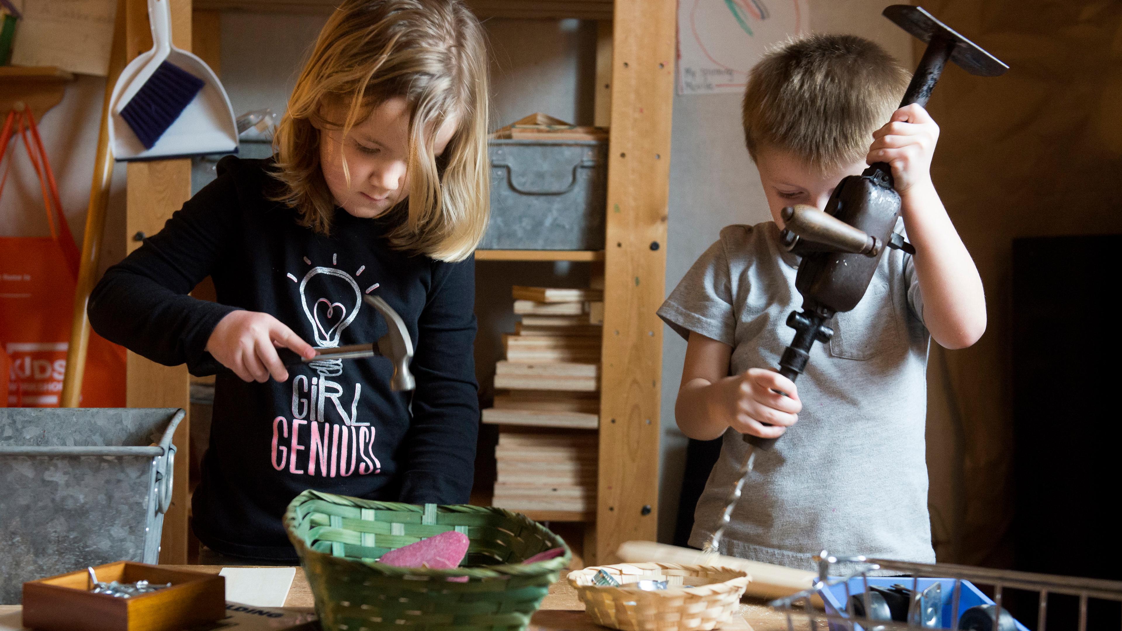 Photo of two children in a workshop setting using tools, one holds a hammer the other uses a hand drill.