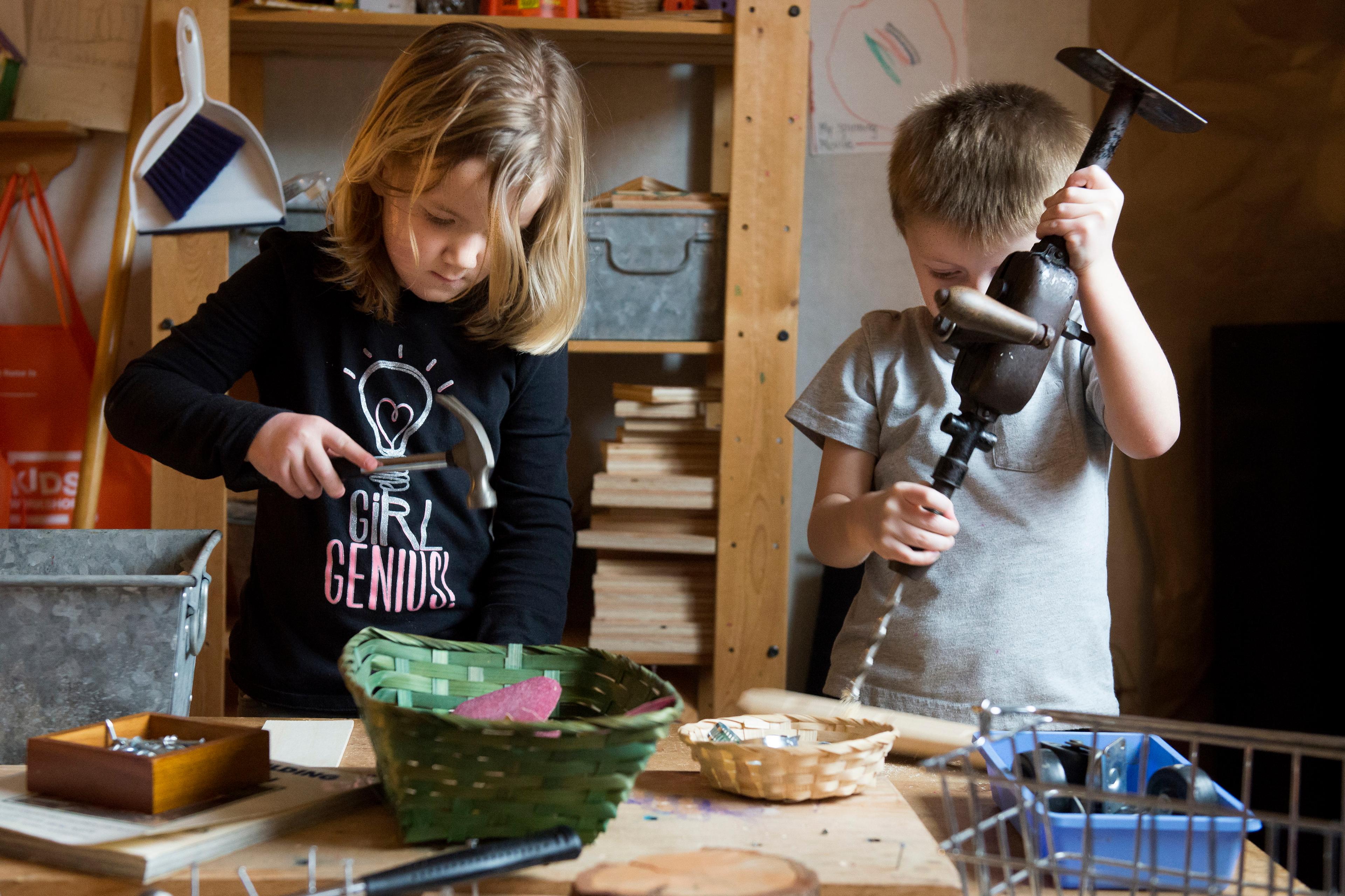 Photo of two children in a workshop setting using tools, one holds a hammer the other uses a hand drill.
