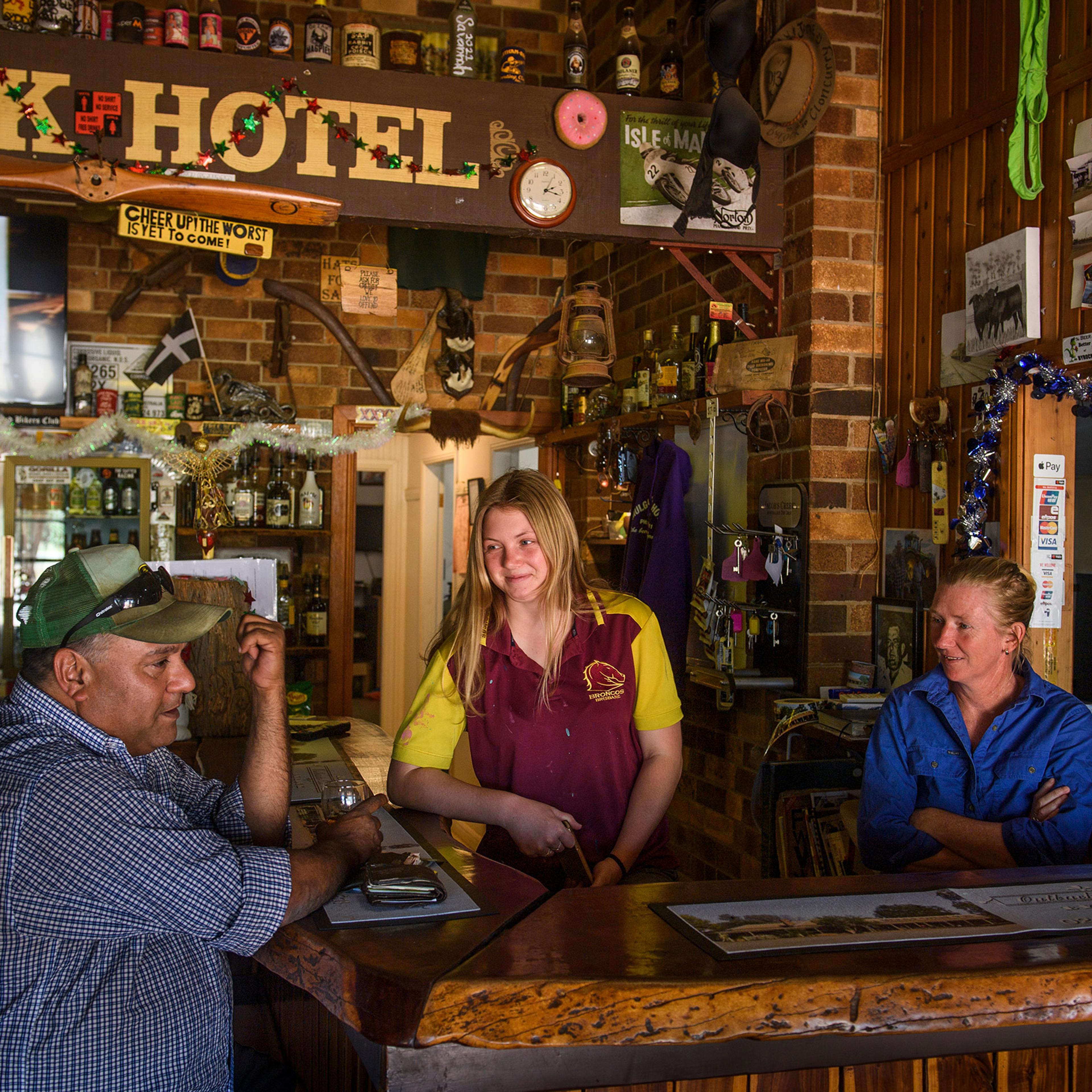 Three people chatting at a rustic bar in a pub, with a brick interior and various decorations.