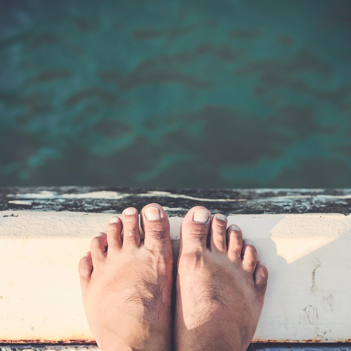 A close up shot looking down upon the toes and feet of a person balanced on the edge of a jetty over water