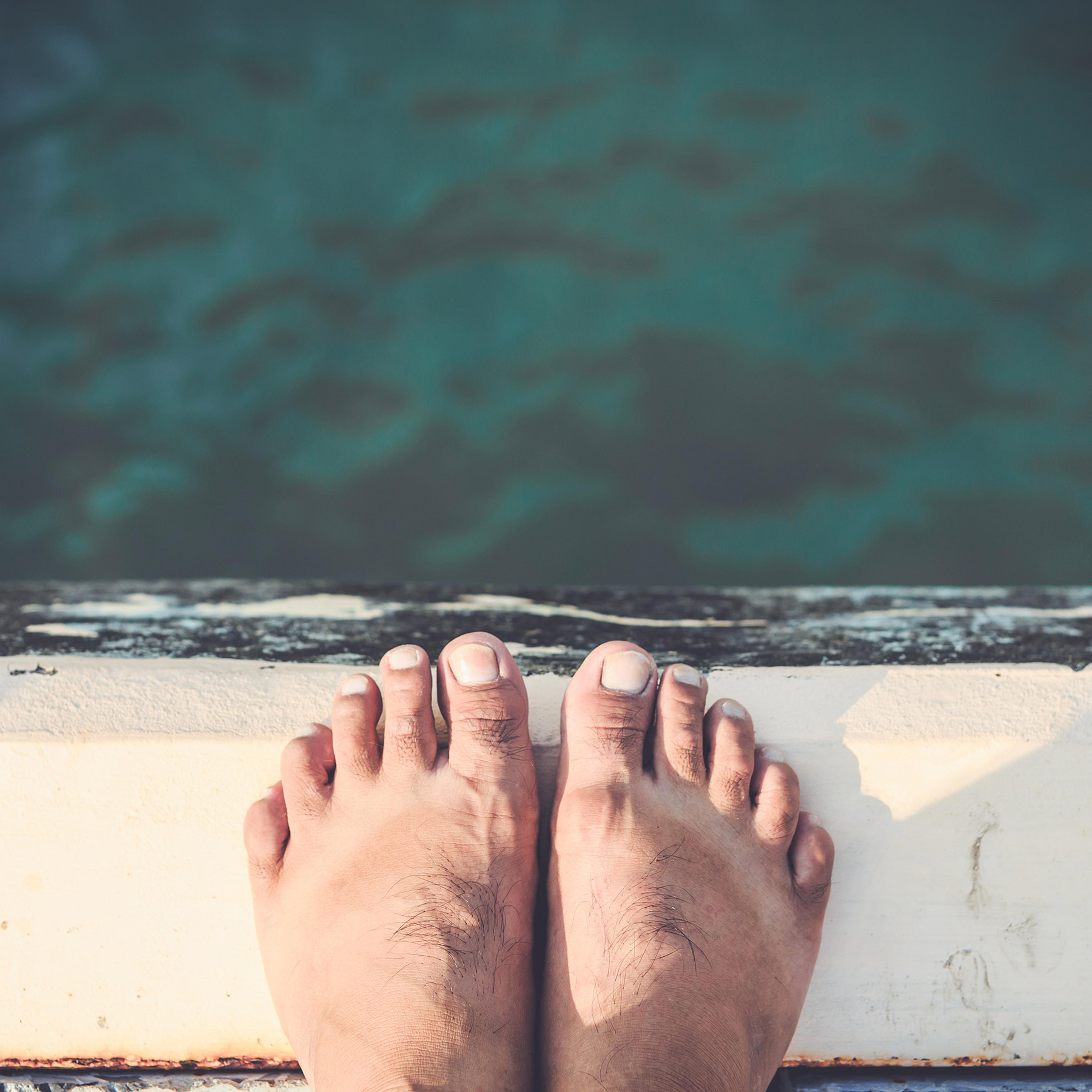 A close up shot looking down upon the toes and feet of a person balanced on the edge of a jetty over water