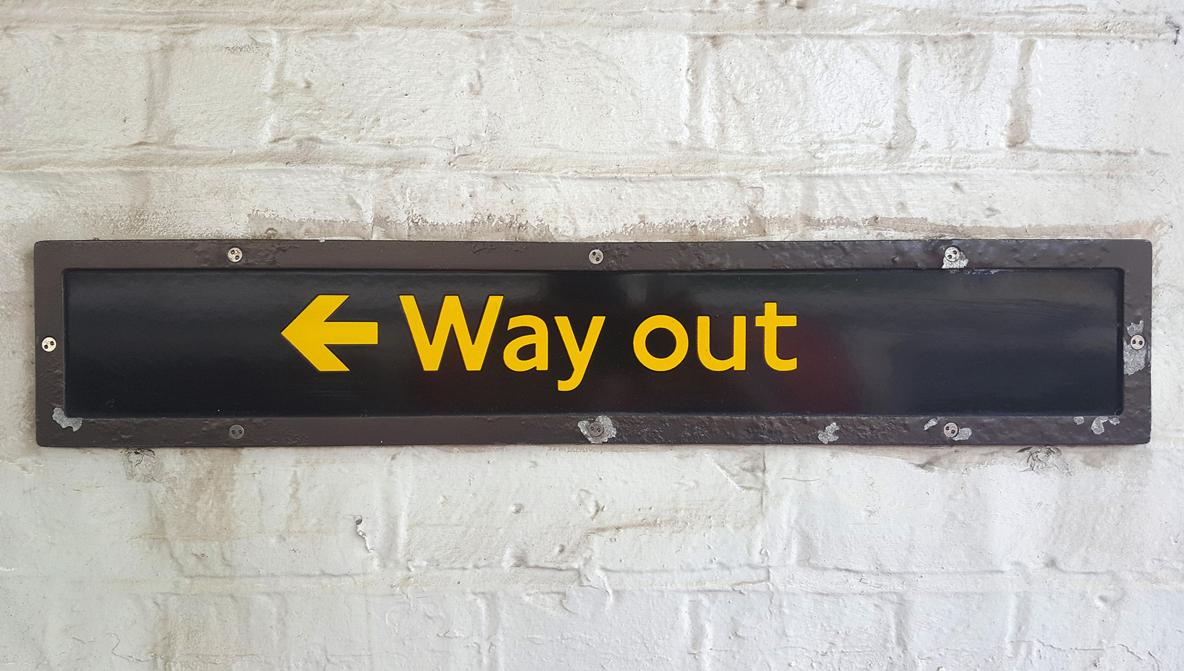 Photo of a black sign with yellow text reading ‘Way out’ and a left-pointing arrow, mounted on a white brick wall.