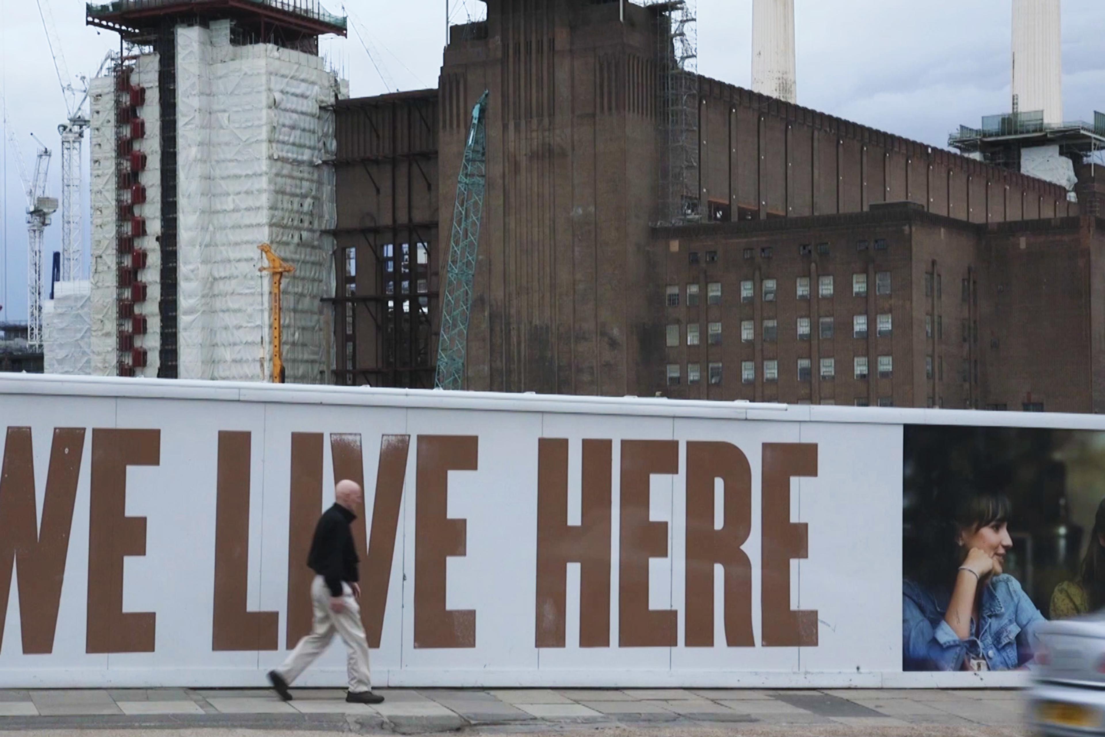 A person walks past a construction site with “WE LIVE HERE” written on a white panel and an image of two women smiling.