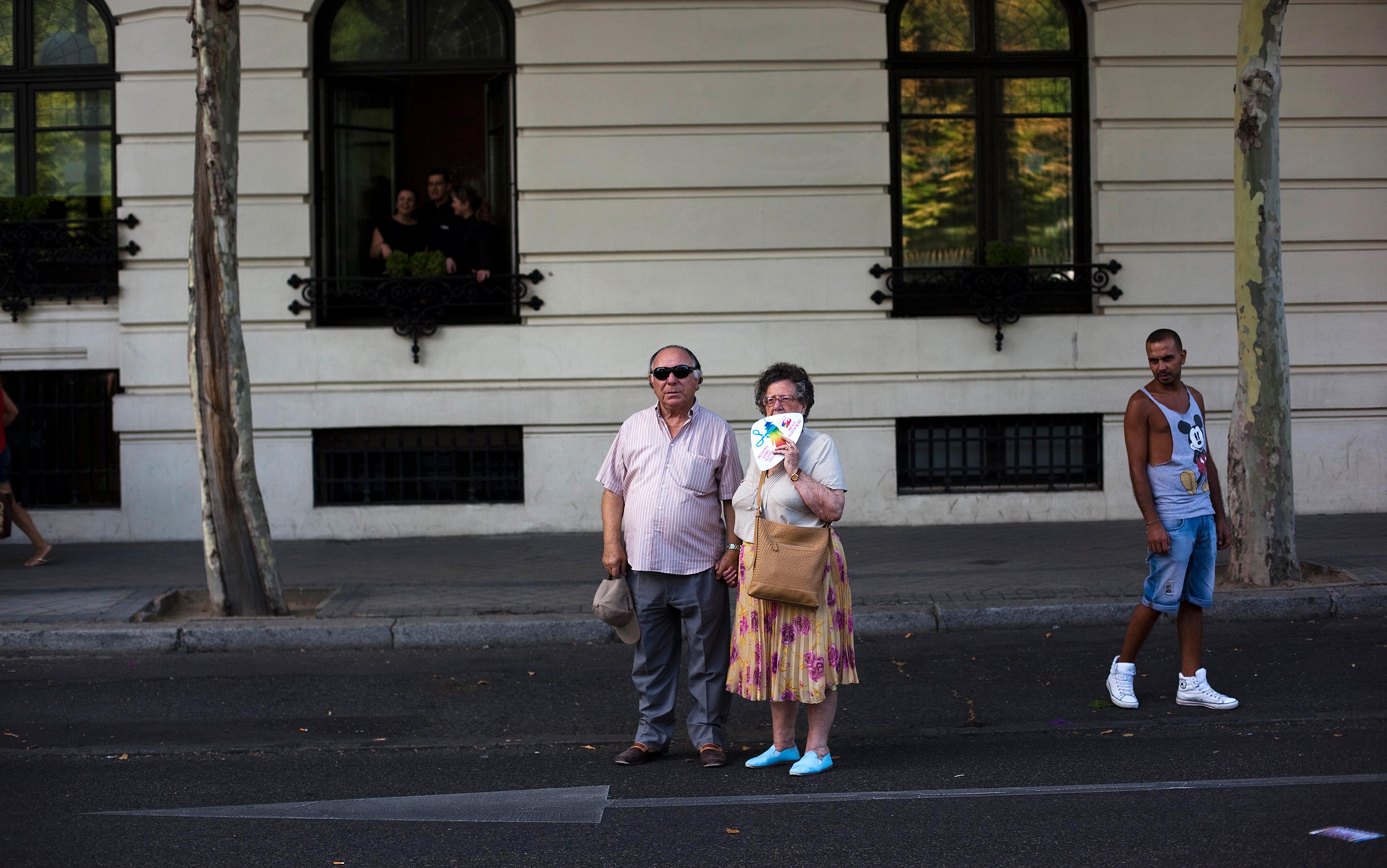 Elderly couple holding hands while standing in the street. The woman holds a colourful fan partially covering her face. A man in casual attire walks by on the right. Two trees and a white building with large windows are in the background, with three people looking out of one of the windows.
