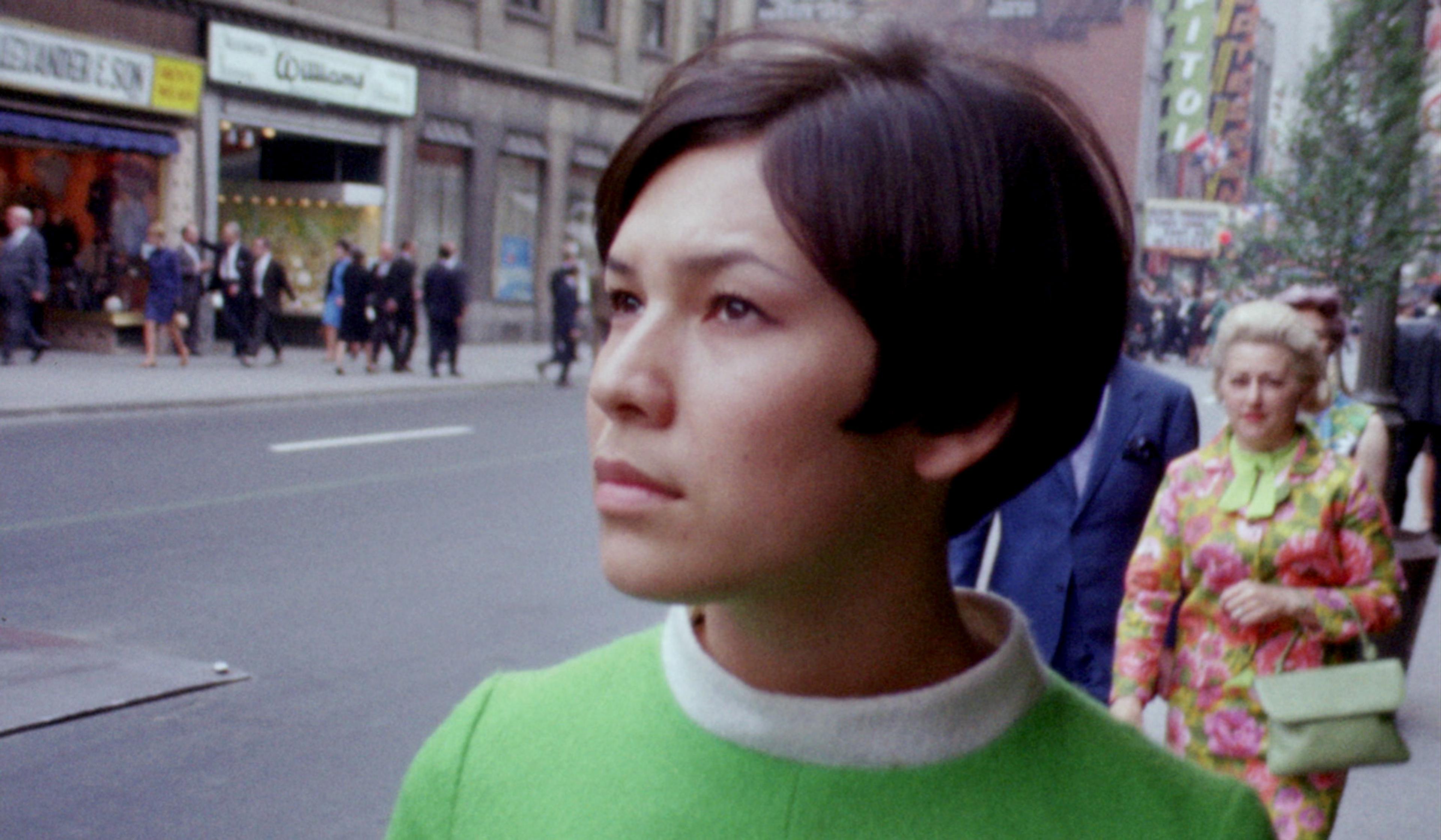 Woman in green top walking down a city street, with shops and other pedestrians in the background.