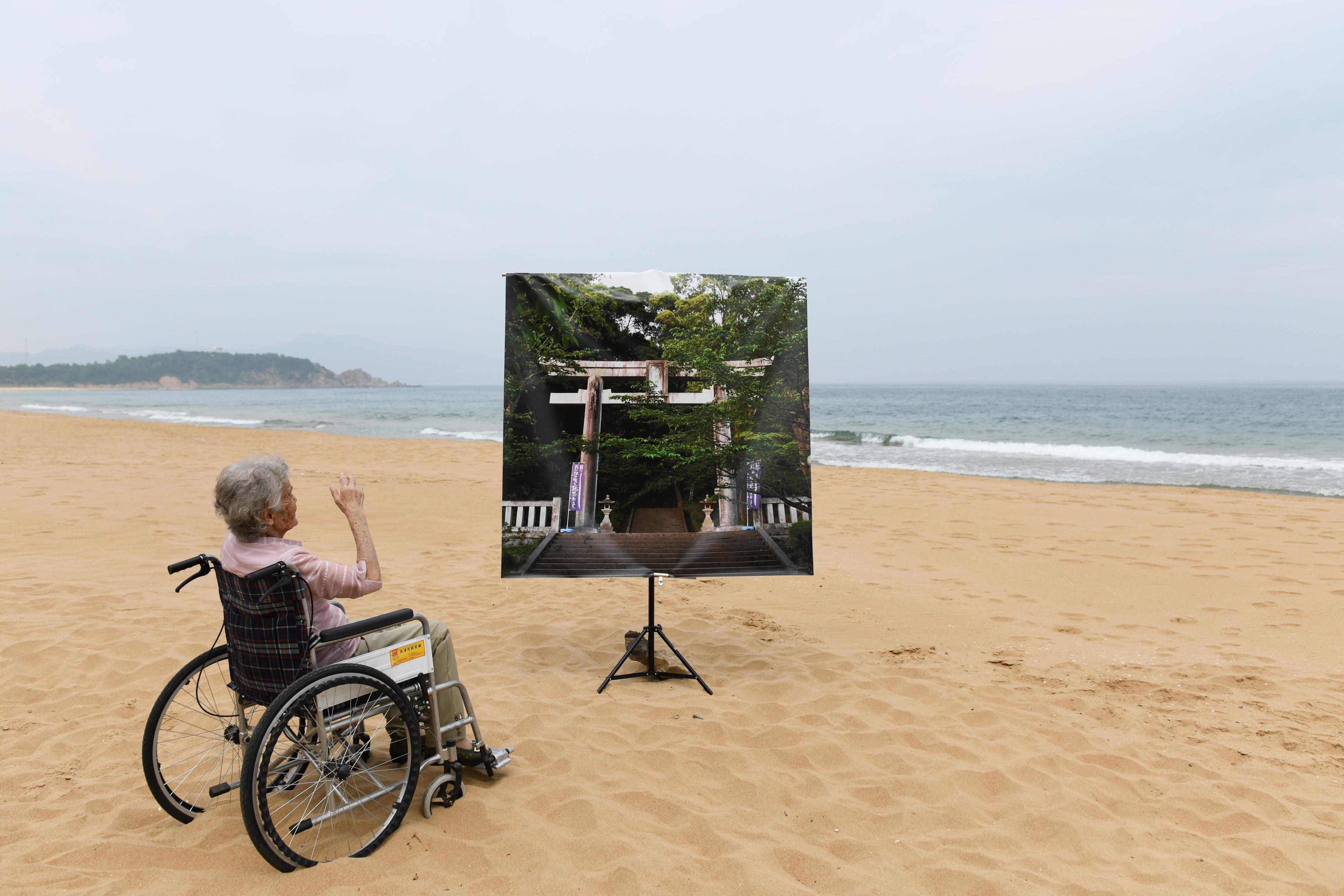 Photo of an elderly person in a wheelchair on a beach facing a large picture of a forest and shrine.