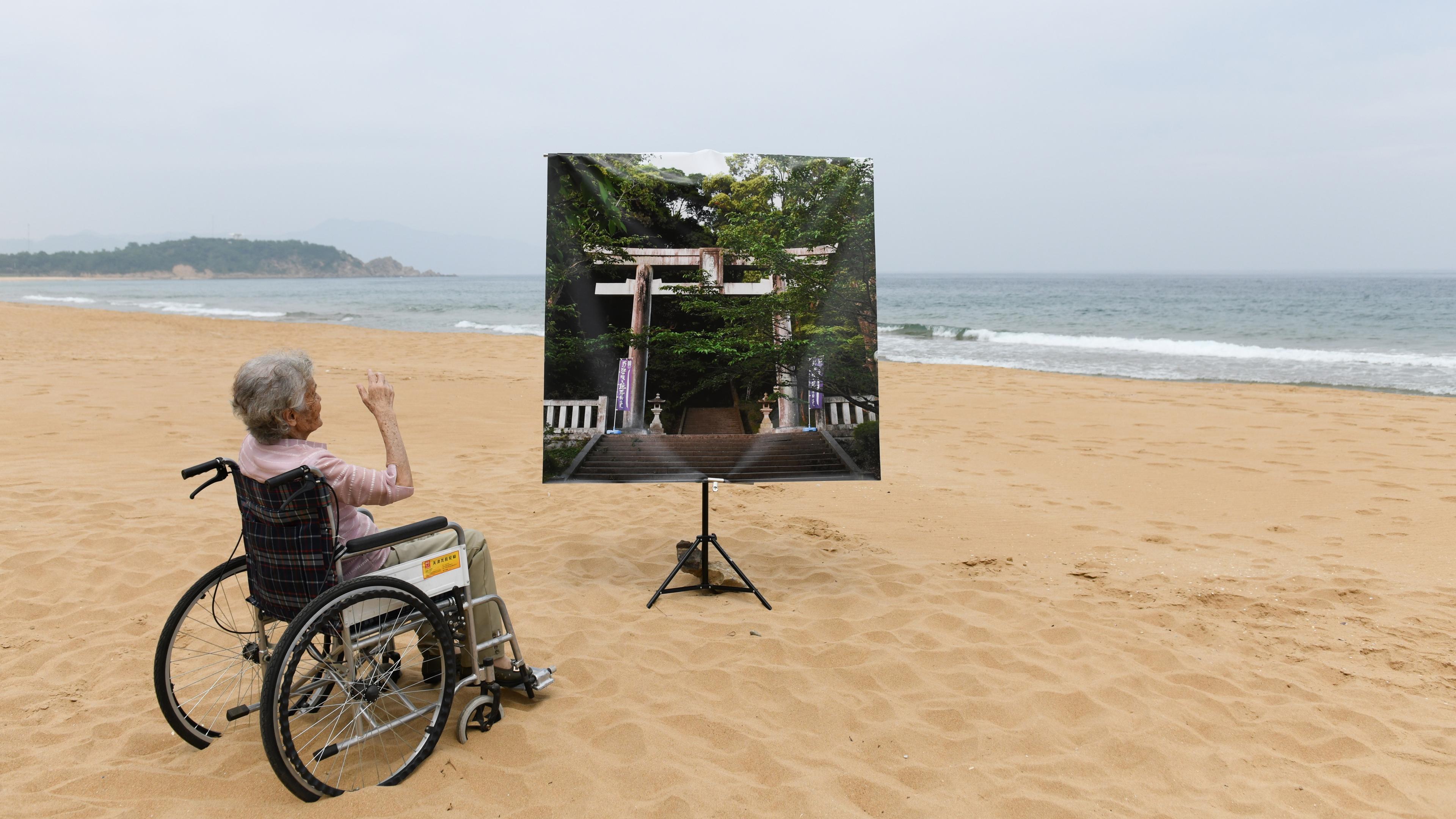 Photo of an elderly person in a wheelchair on a beach facing a large picture of a forest and shrine.