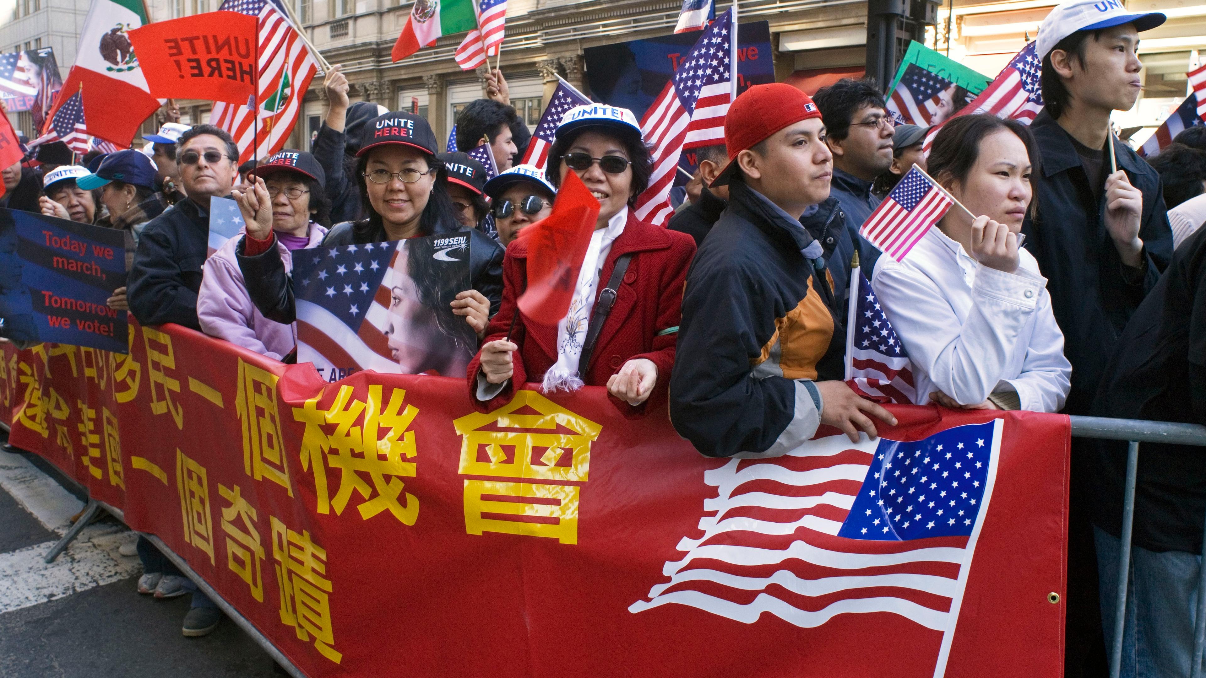 Photo of a diverse crowd holding American flags and banners during a street protest, with buildings in the background.