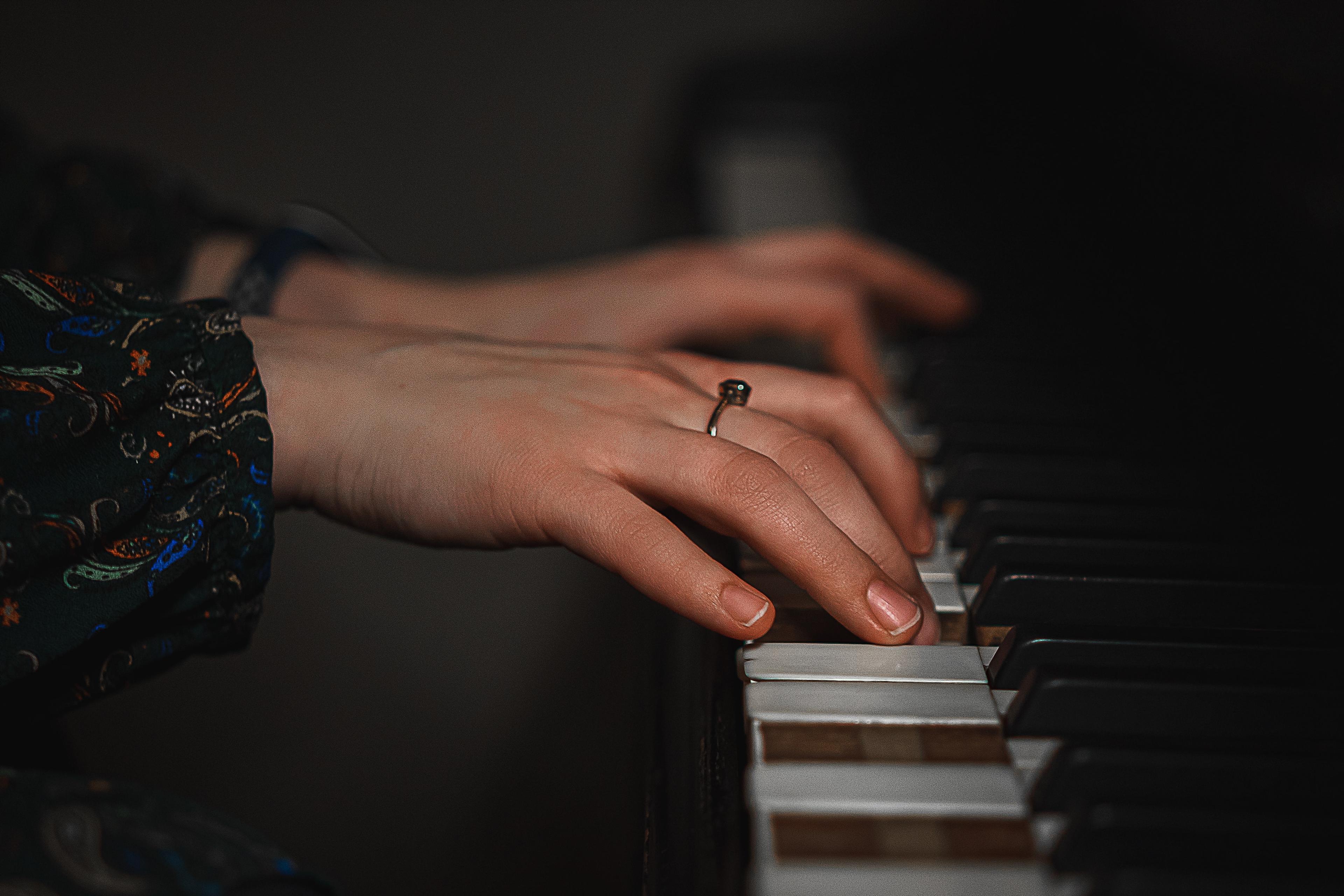 Photo of hands playing a piano keyboard, showing close-up of fingers and a ring on one finger against dark background.