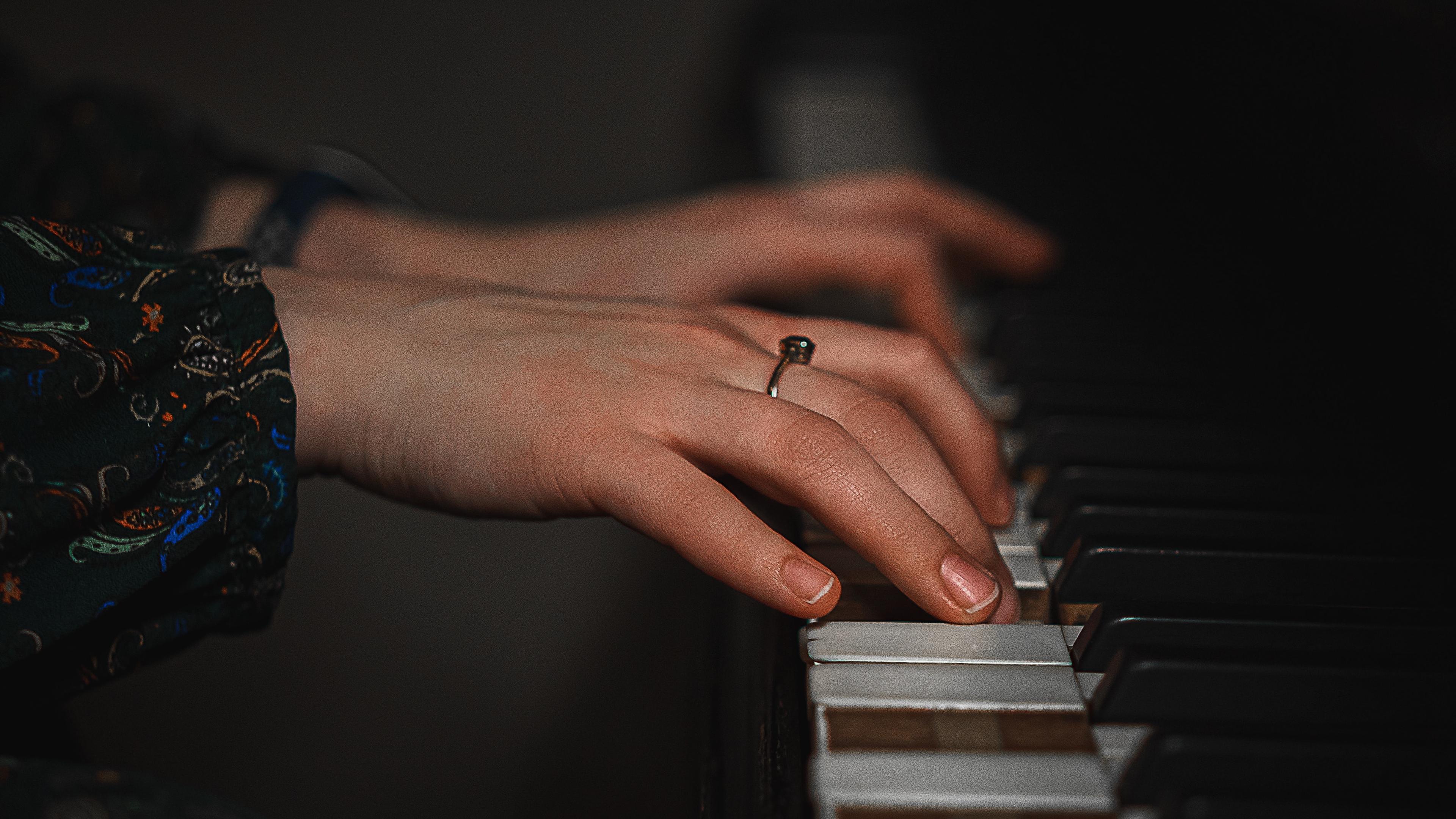 Photo of hands playing a piano keyboard, showing close-up of fingers and a ring on one finger against dark background.