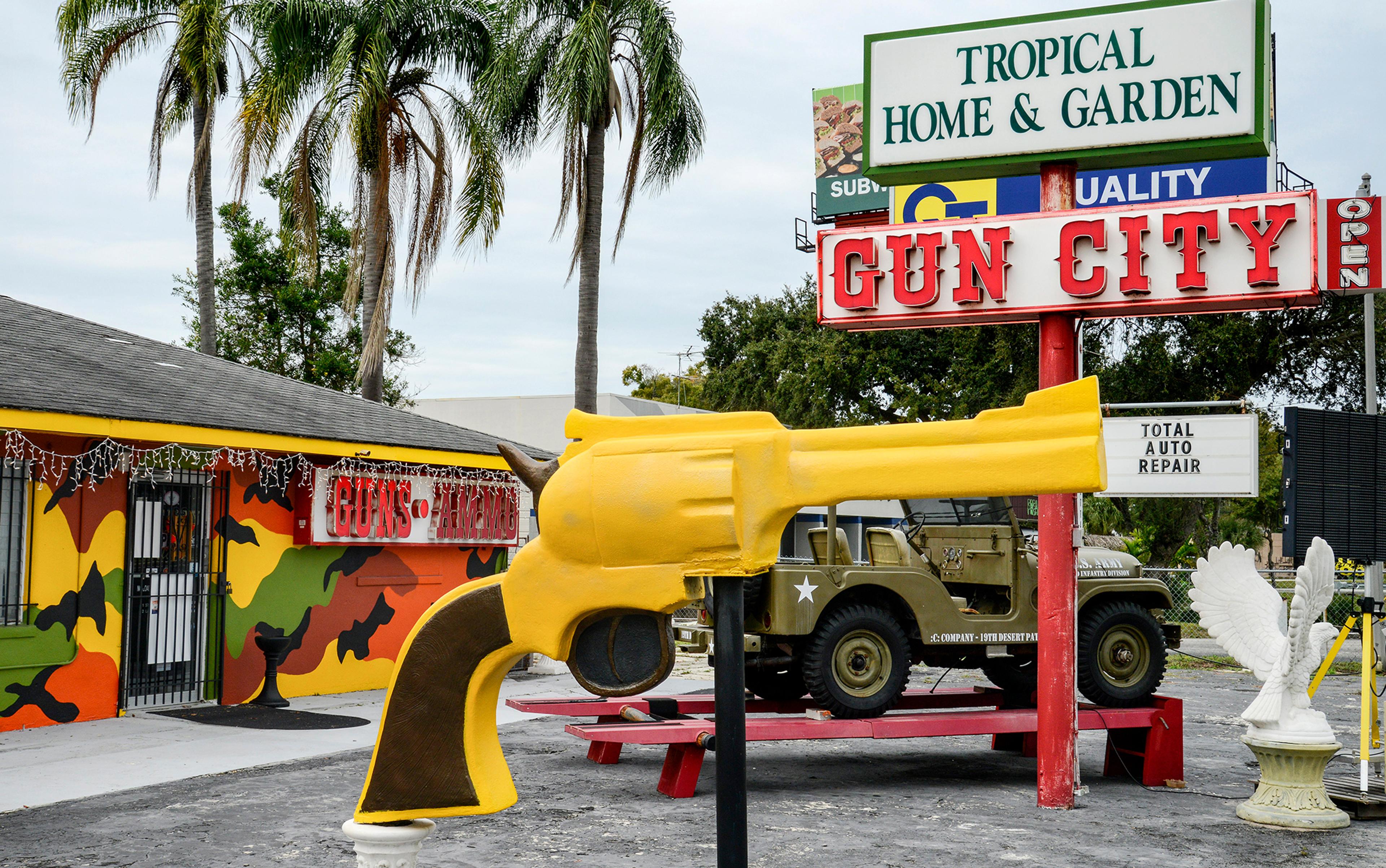 An oversized yellow plastic revolver gun displayed outside a brightly painted shop. There are palm trees in the background