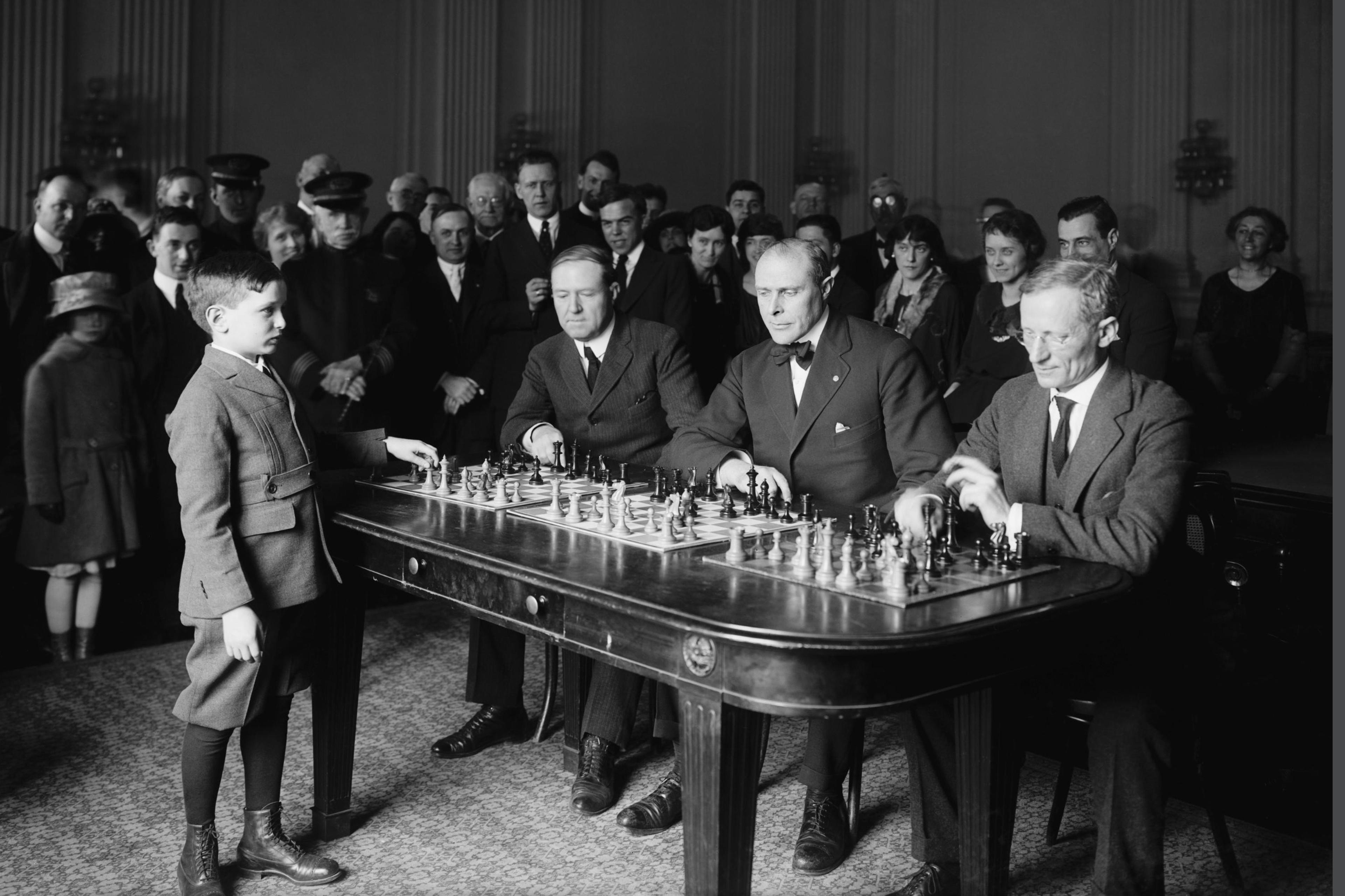 Black and white photo of a young boy playing simultaneous chess against three suited men watched by a crowd.