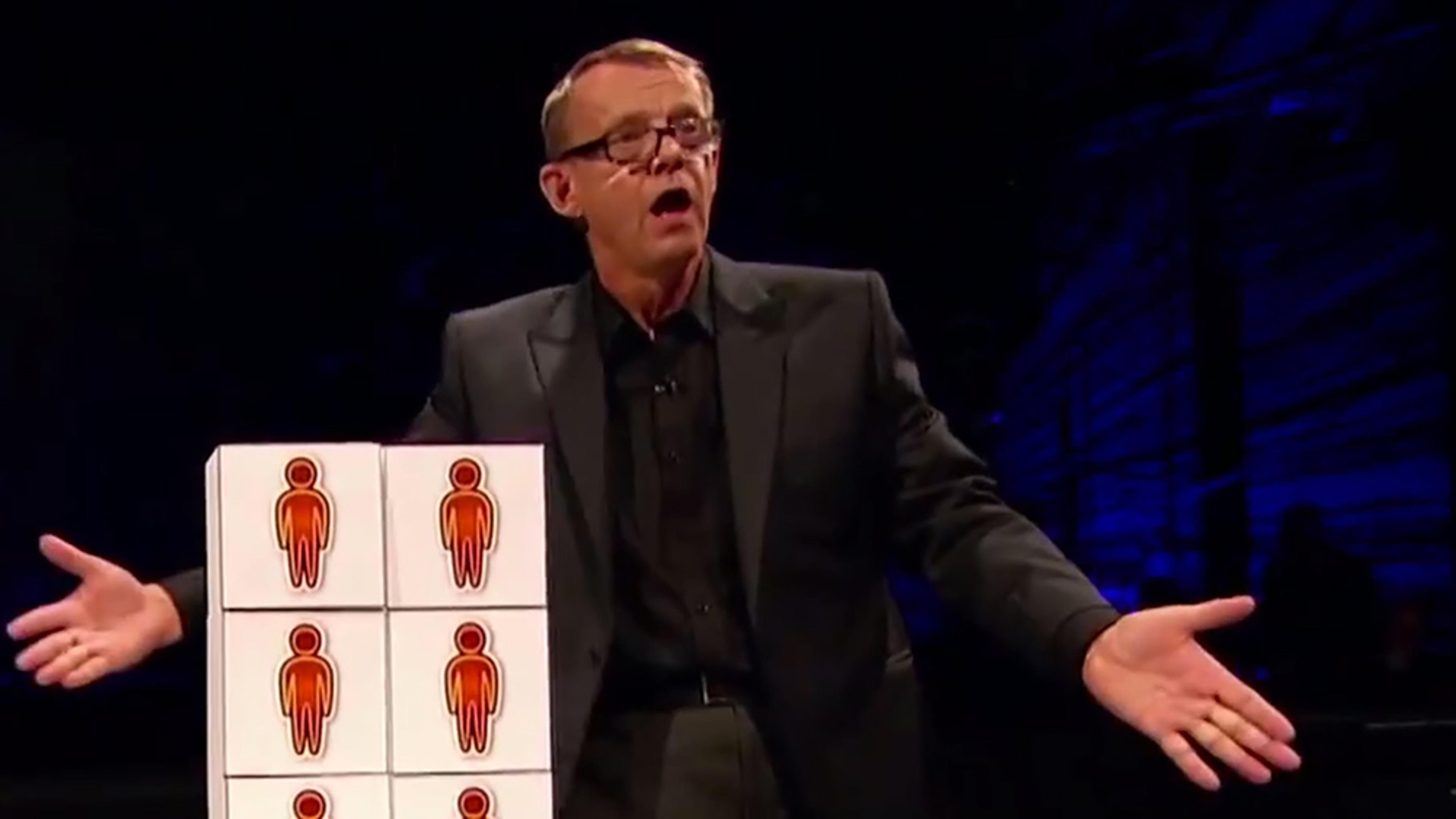 A man gestures while standing next to a set of blocks with red human figures in a dimly lit environment.