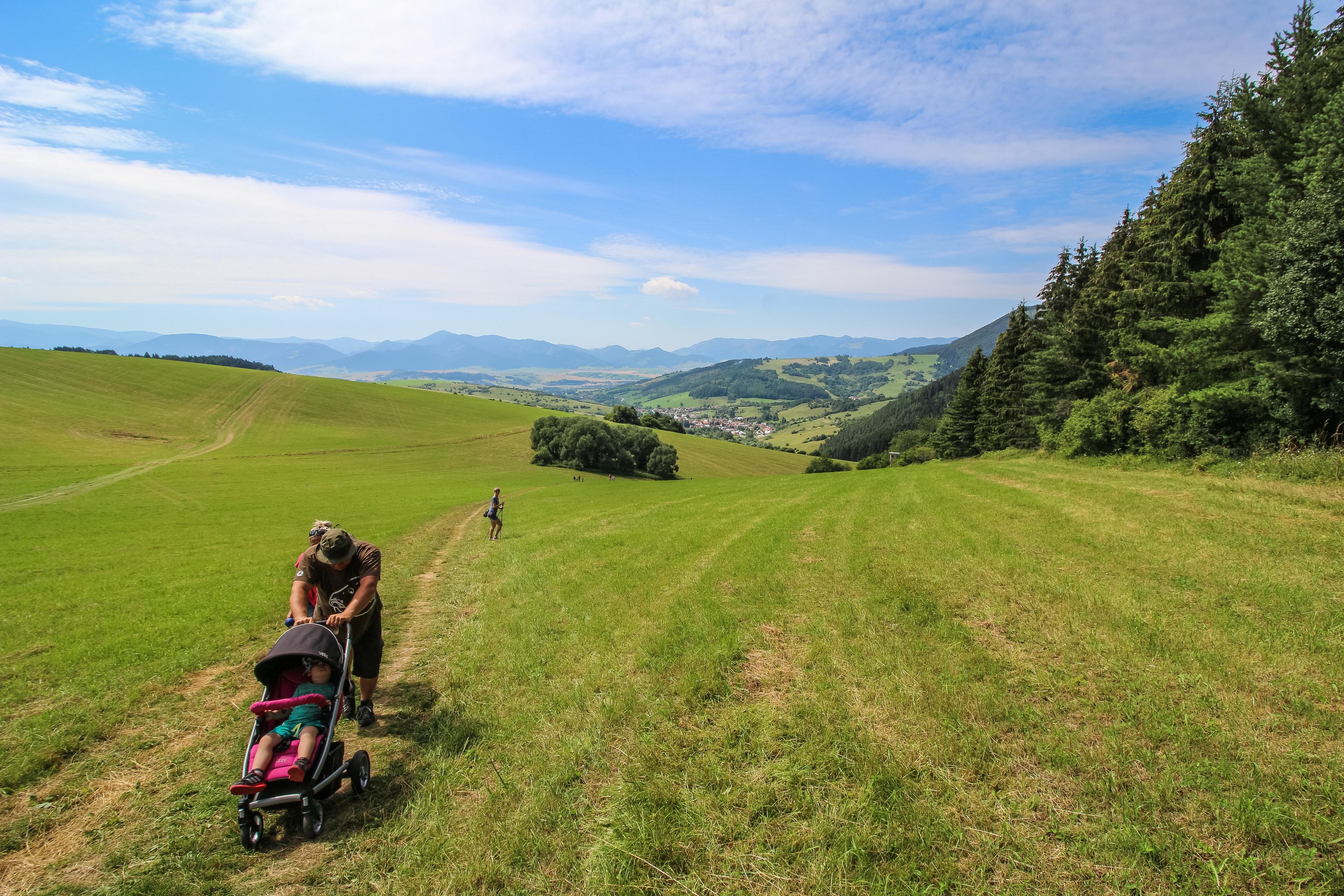 Photo of a person pushing a pram on a grassy hill with mountains and a village in the background.