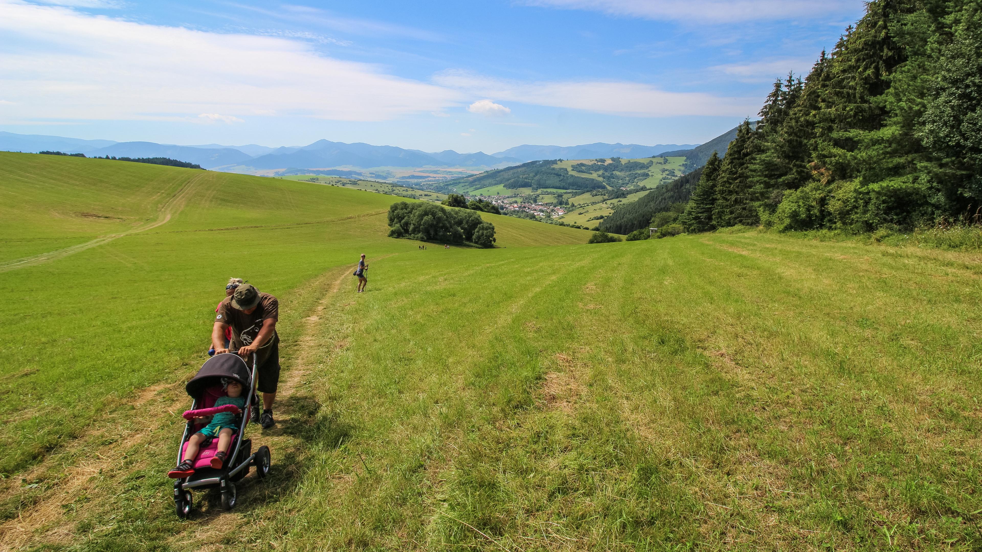 Photo of a person pushing a pram on a grassy hill with mountains and a village in the background.