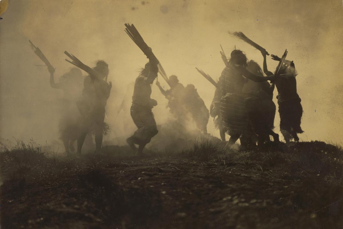 Sepia-toned photo of silhouetted figures dancing with sticks in a smoky environment, creating a dramatic atmosphere.