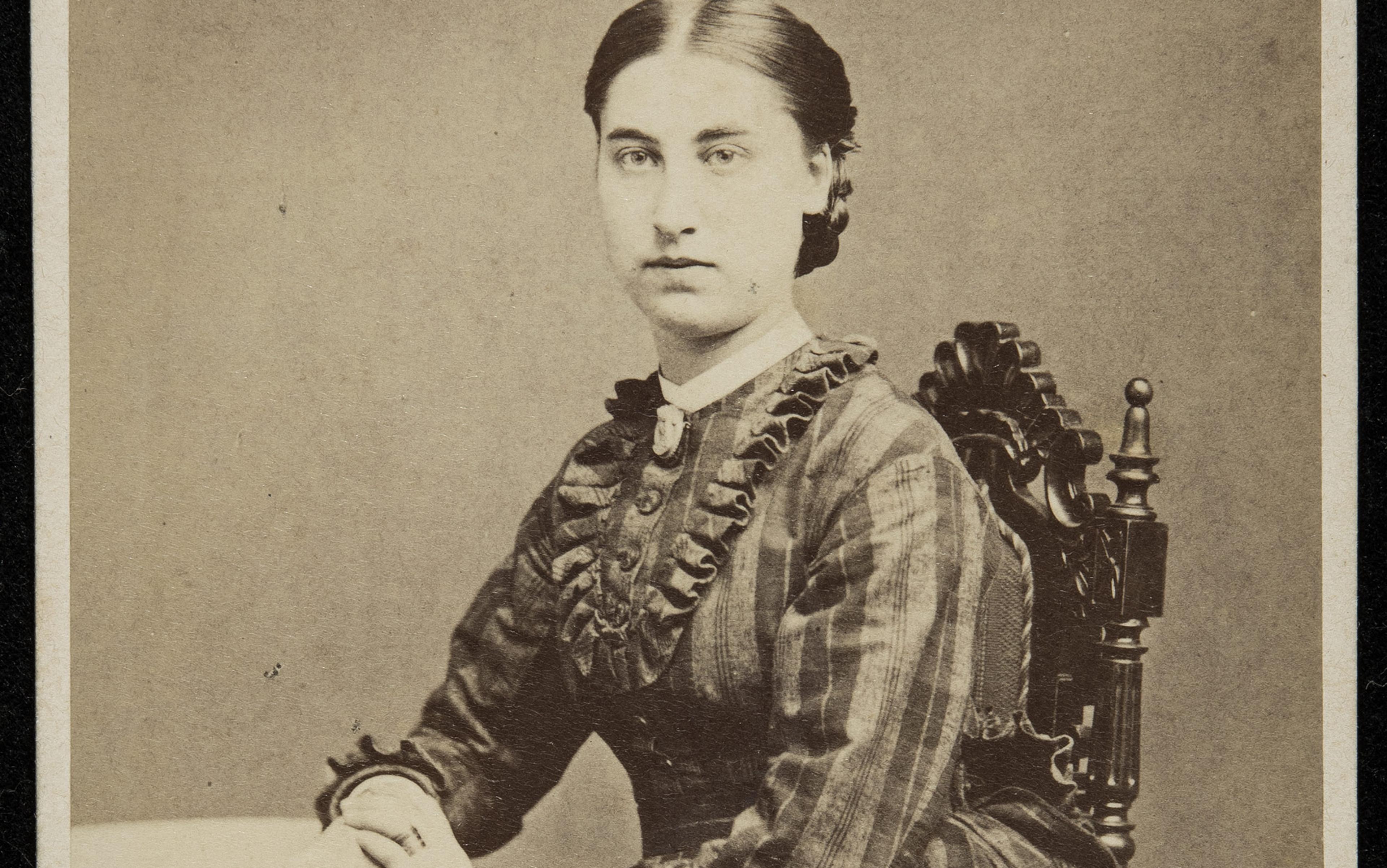 A sepia photograph of a woman from the 19th century, sitting on an ornate wooden chair, wearing a ruffled dress with buttons.
