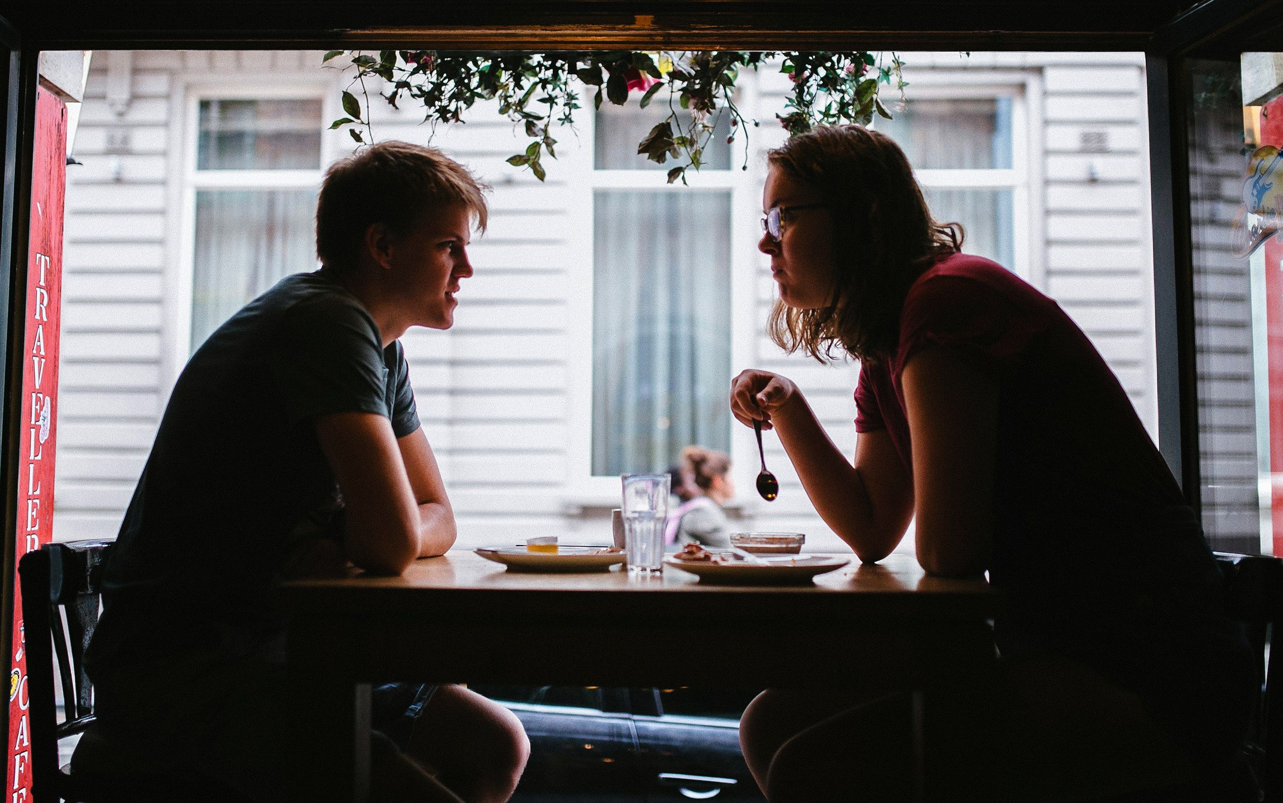 Two people sitting at a cafe table, silhouetted against a window with hanging plants above.