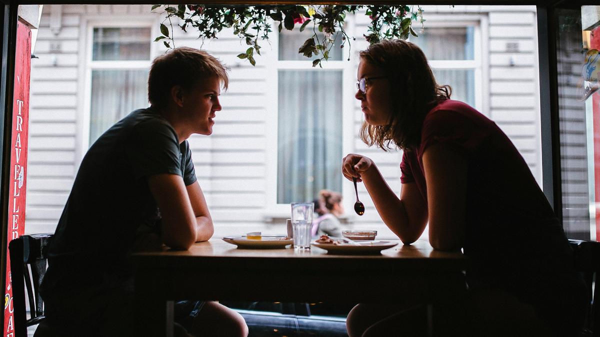 Two people sitting at a cafe table, silhouetted against a window with hanging plants above.