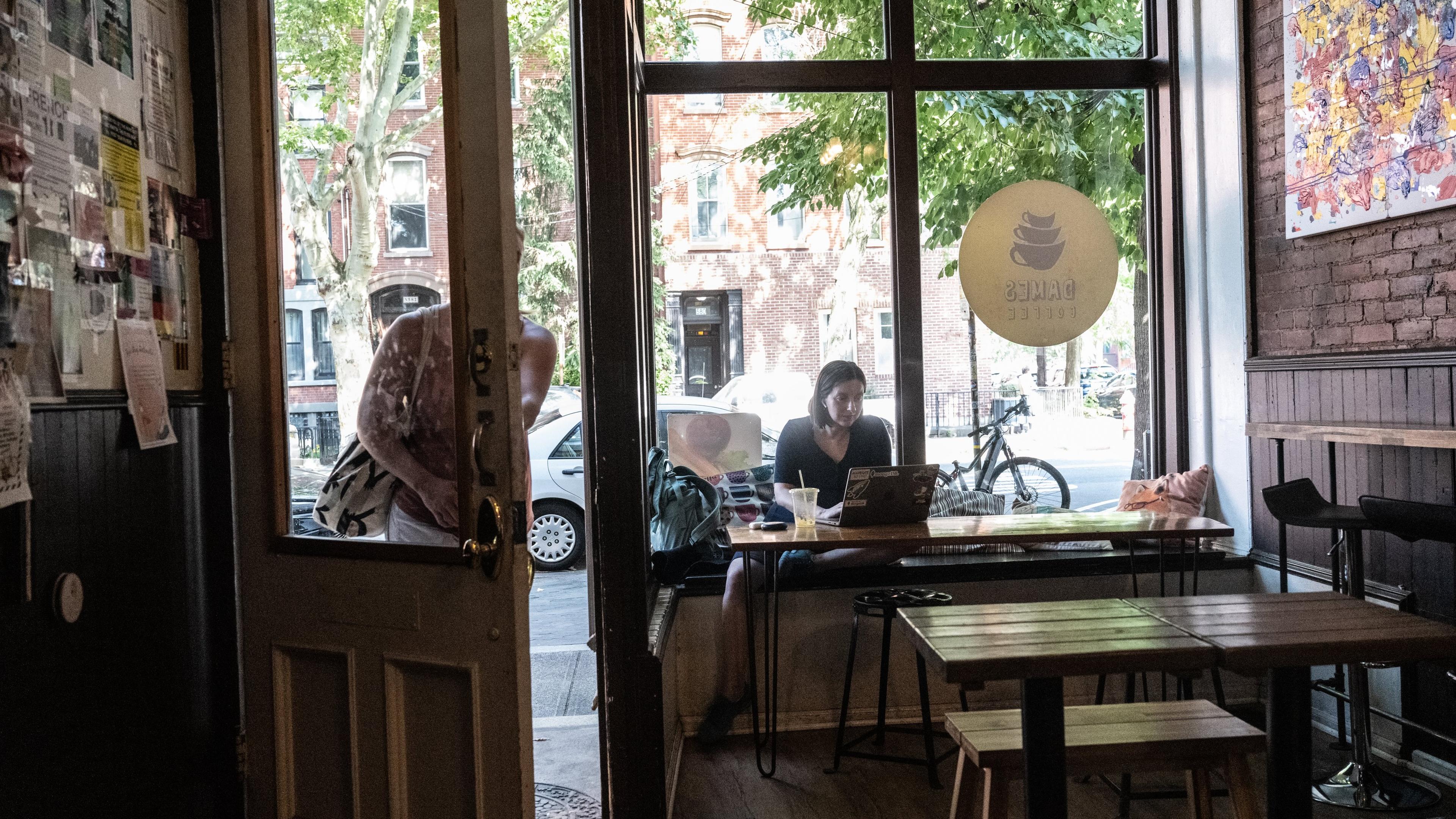 Photo of a cafe interior with a person working on a laptop by a window, open door and street view with bicycle outside.