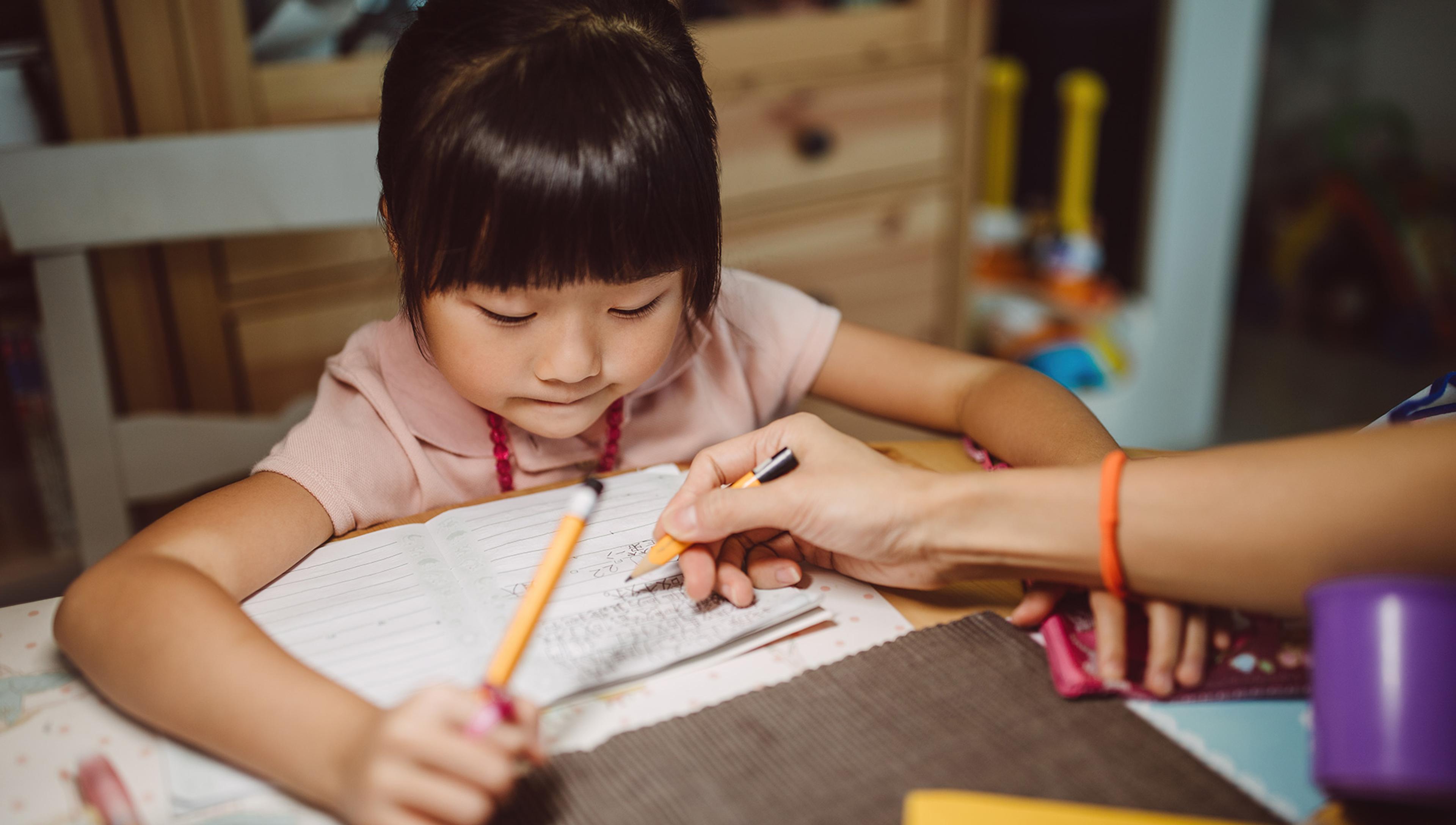 Photo of a child in a pink shirt concentrating on writing in a notebook while an adult assists, showing a learning activity.