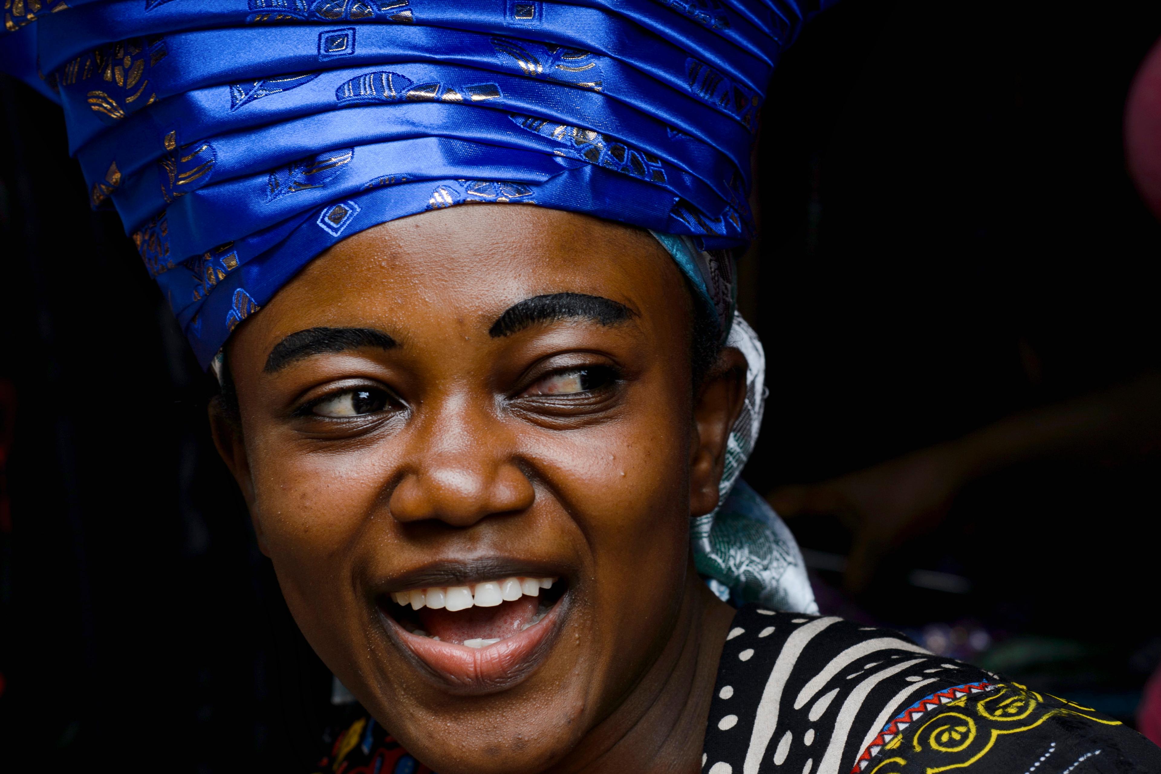 Photo of a smiling person in vibrant attire and a large blue head wrap against a dark background.