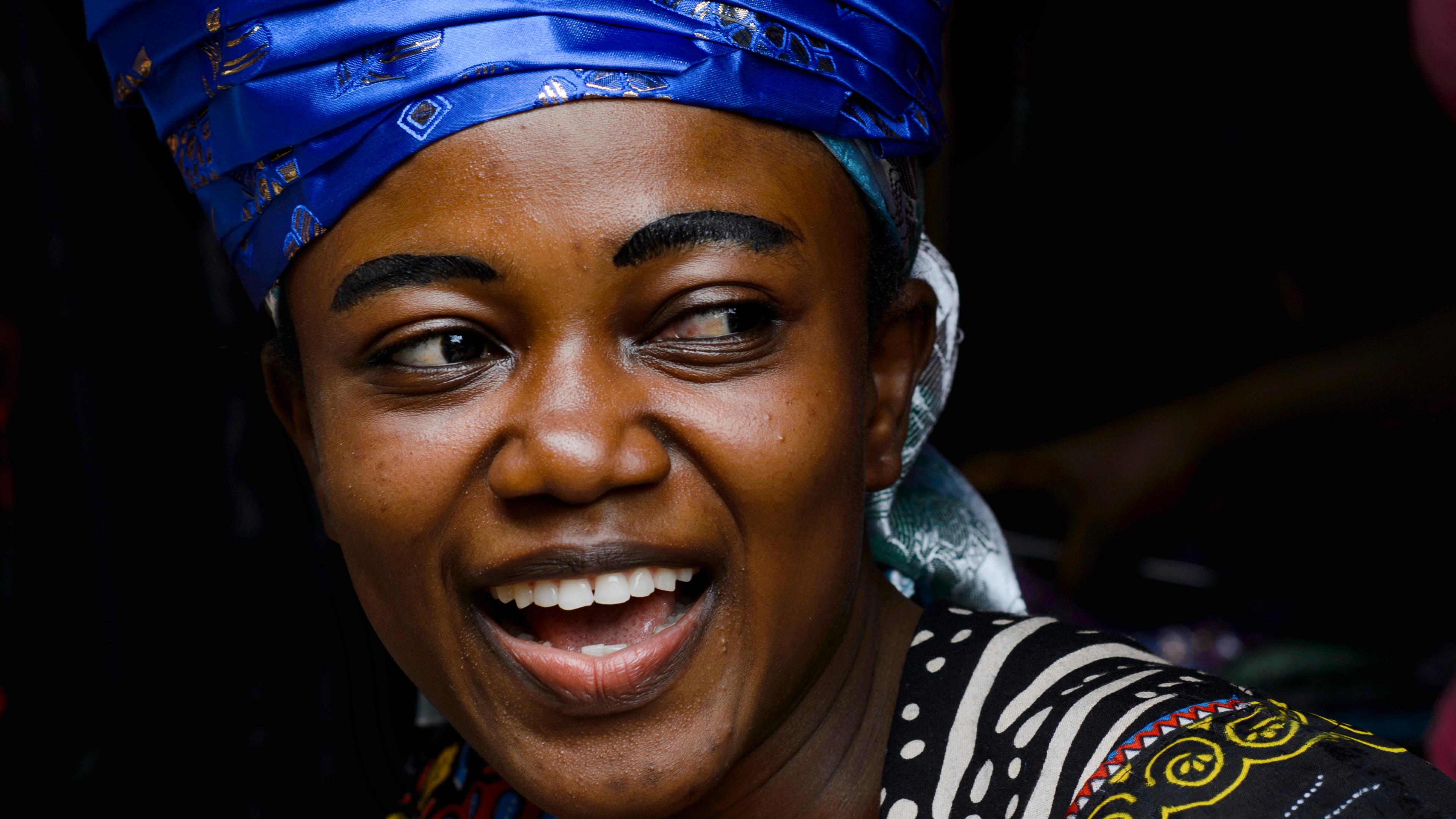 Photo of a smiling person in vibrant attire and a large blue head wrap against a dark background.