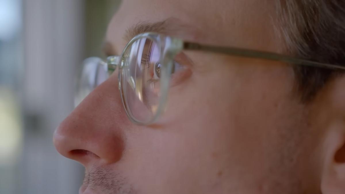 Close-up photo of a person’s face in profile wearing glasses looking out of a window, with reflections in the lenses.