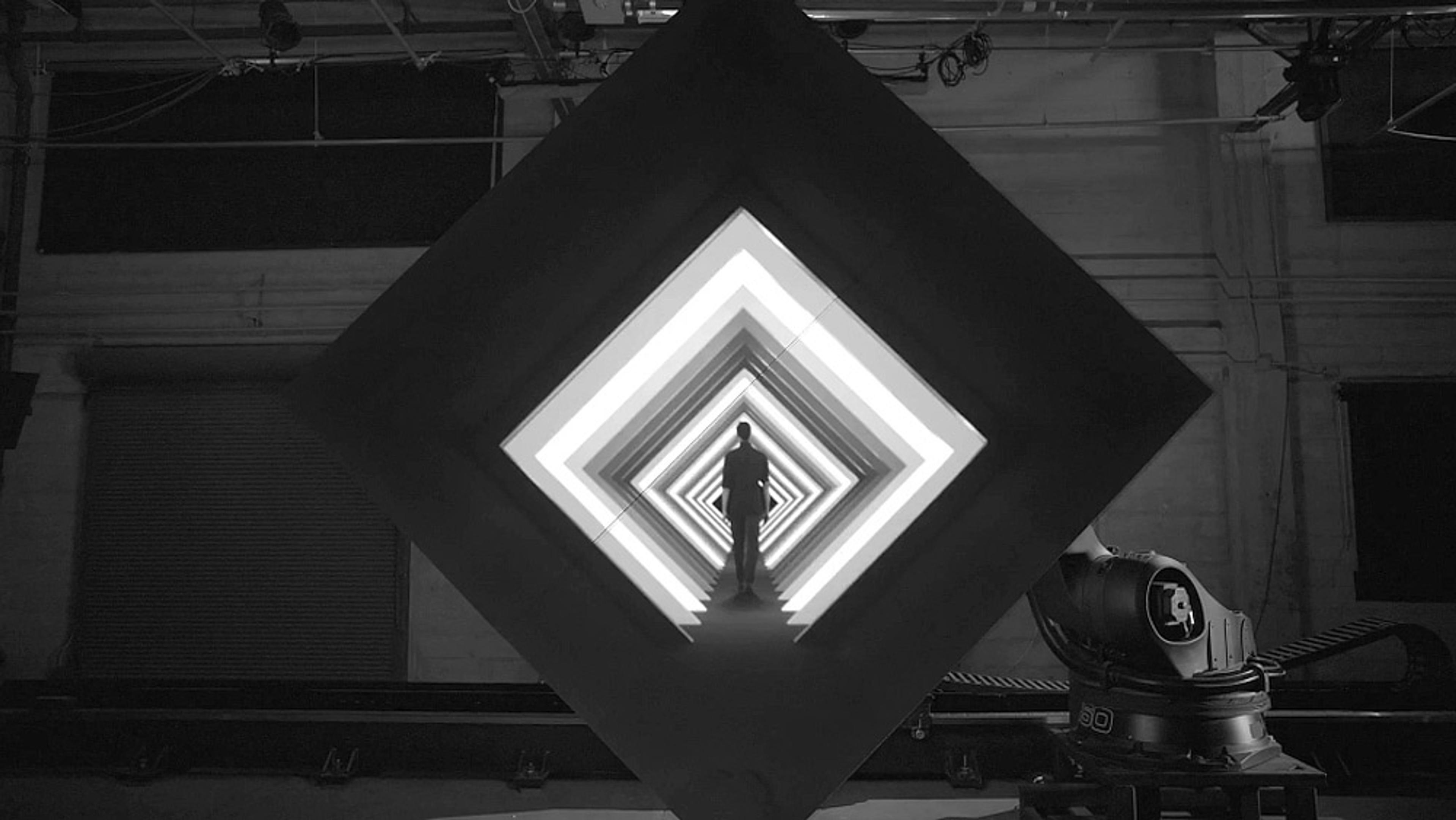 Black and white photo of a person standing inside a square tunnel with illuminated concentric squares, creating a geometric pattern.