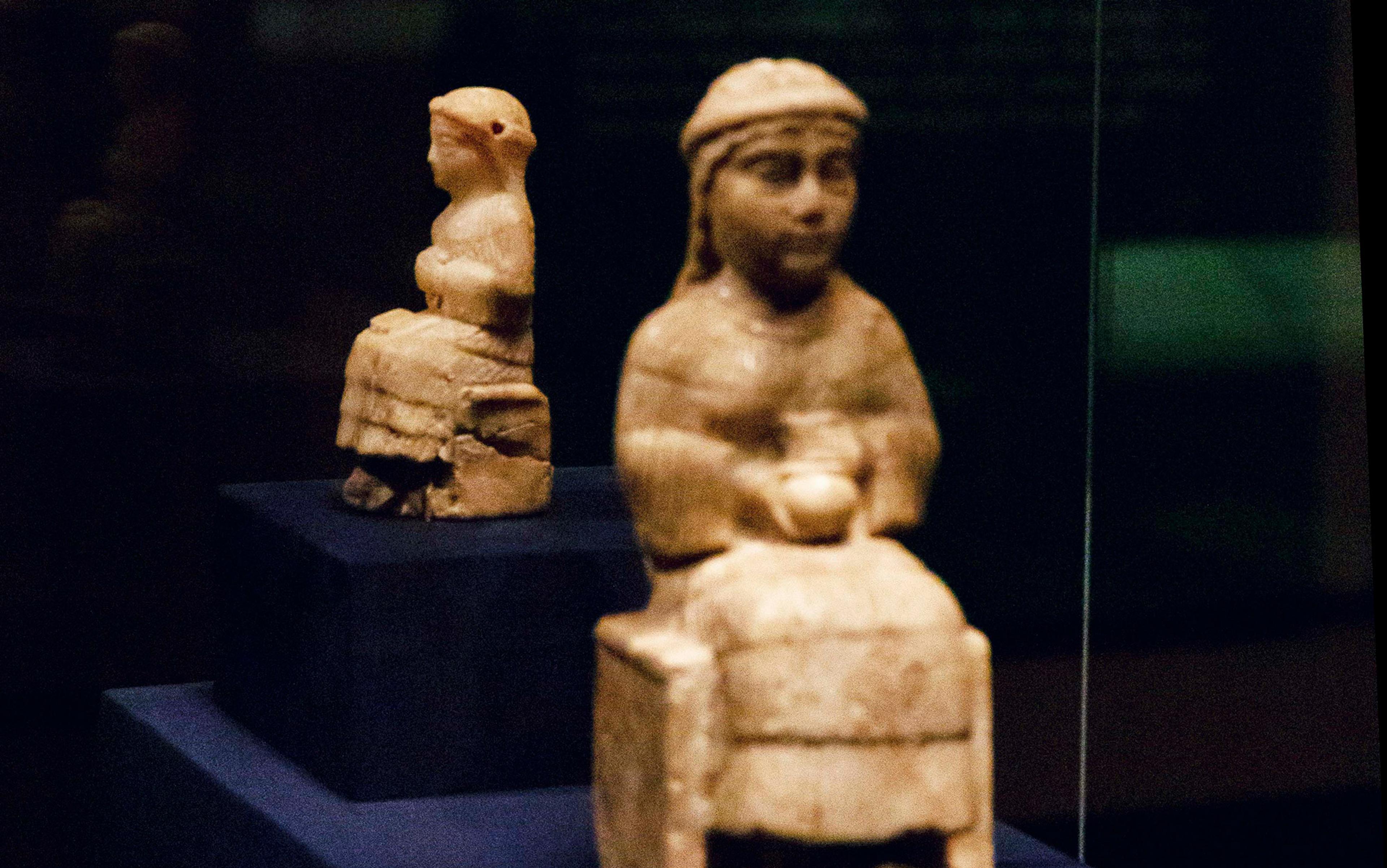 Two small ancient stone statues, each depicting a seated figure, displayed on dark stands in a dimly lit setting.