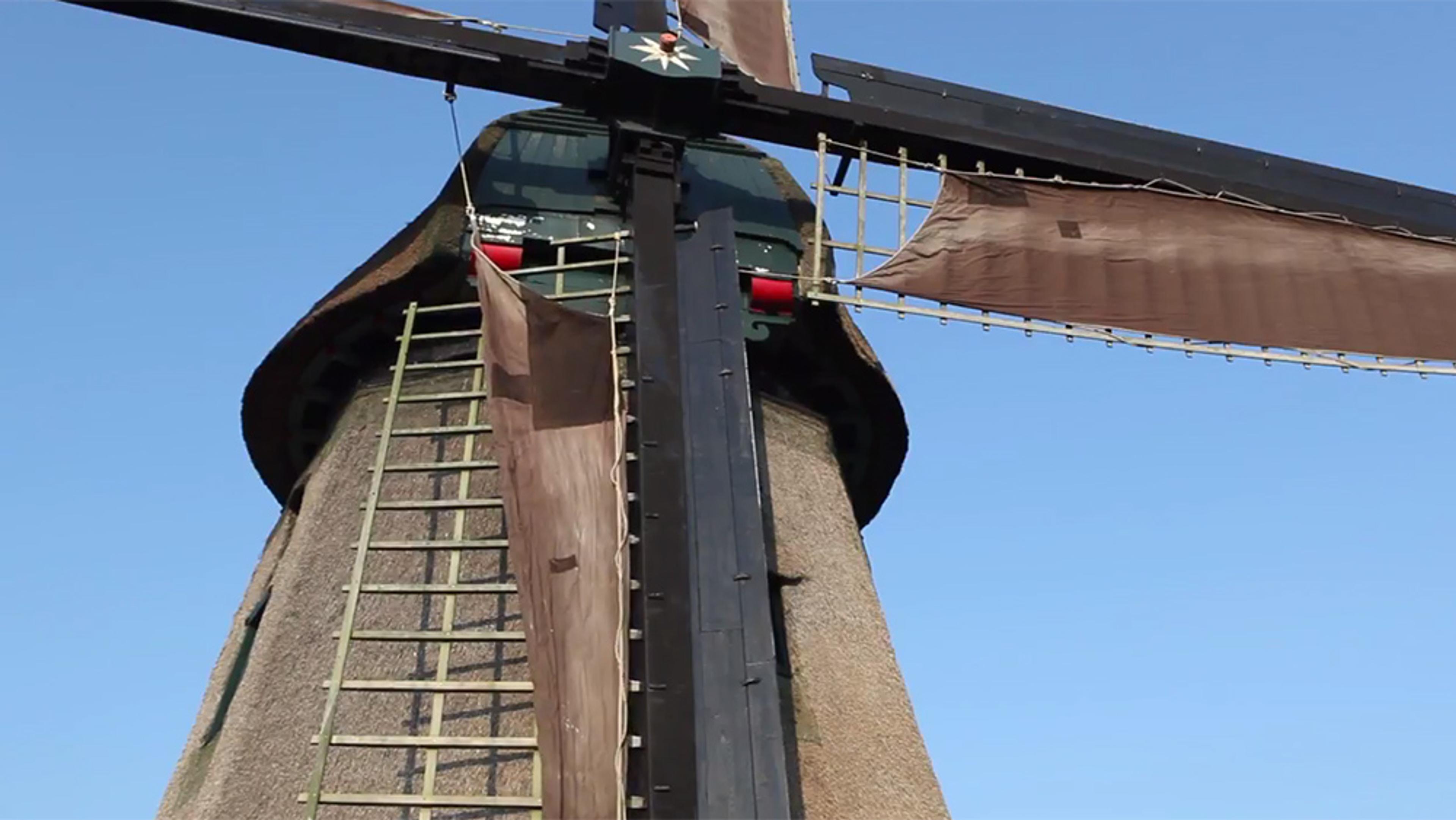 A traditional windmill with large wooden blades against a clear blue sky, focusing on the central structure and one blade.