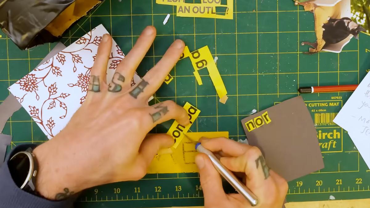 Photo of tattooed hands cutting yellow paper with a craft knife on a green cutting mat scattered with paper pieces.