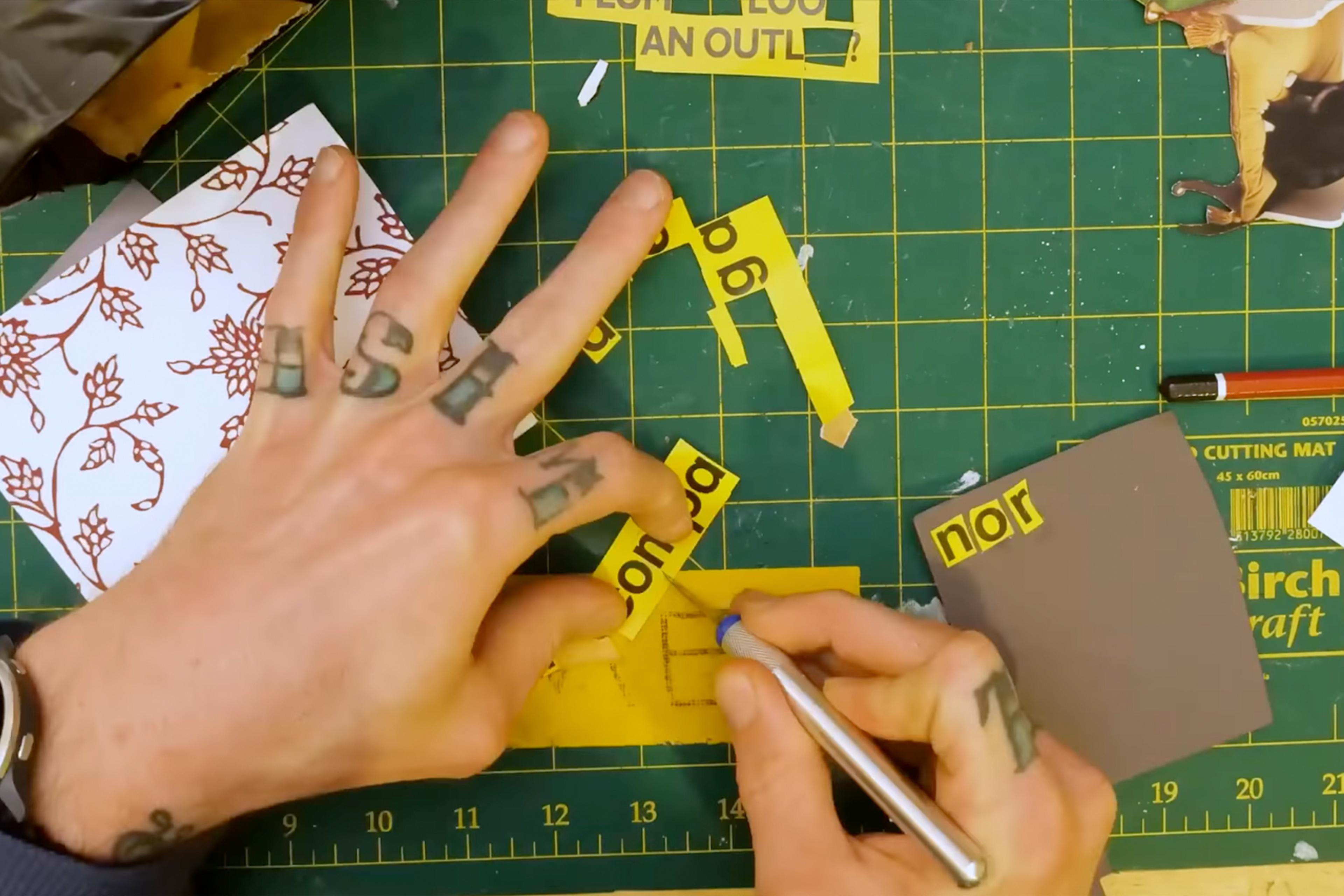 Photo of tattooed hands cutting yellow paper with a craft knife on a green cutting mat scattered with paper pieces.