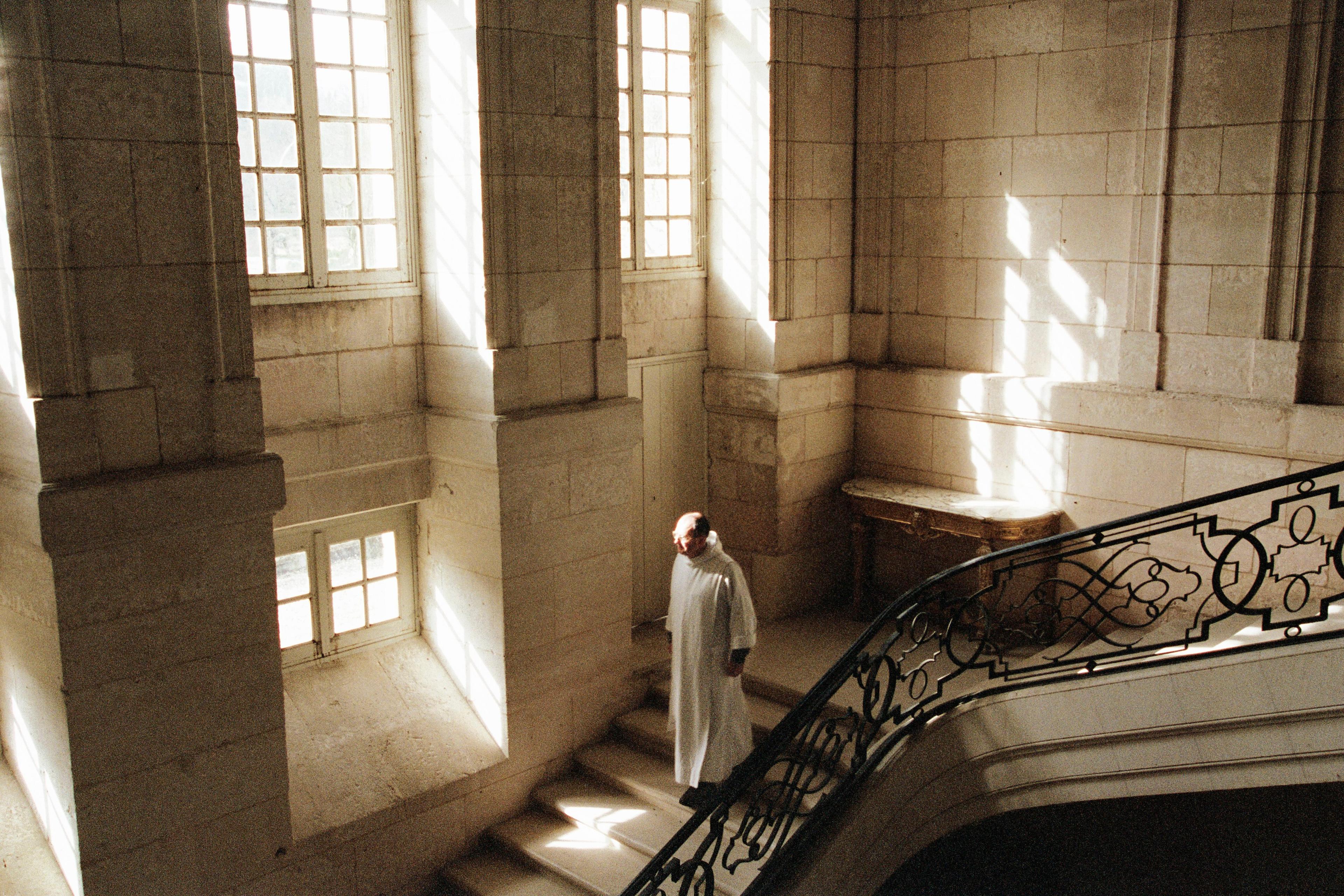 Photo of a person in a white robe on a sunlit stone staircase with large windows and ornate railings.
