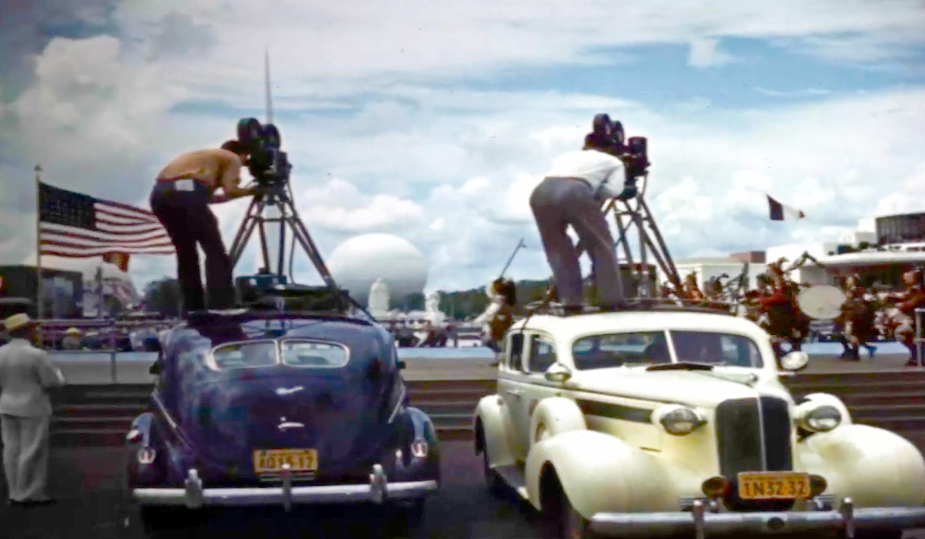 Two men on top of vintage cars filming with large cameras, a US flag, and a marching band in the background.