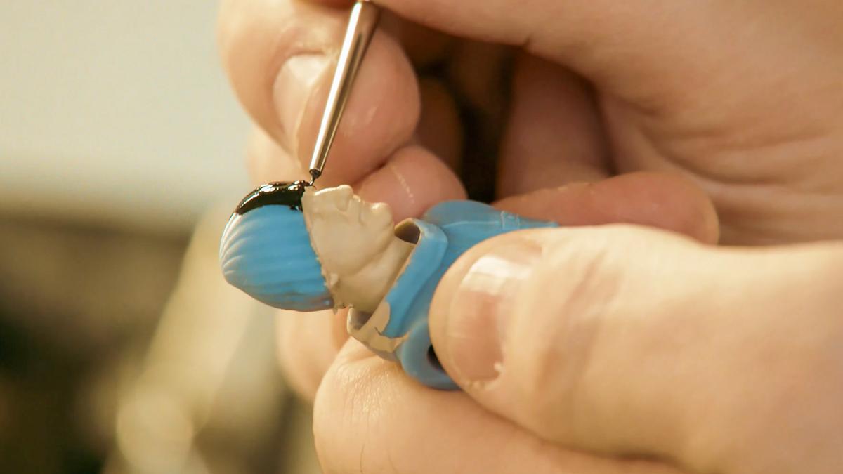 Hands intricately painting a small figurine’s hair black with a fine brush, the figure wears a blue outfit.