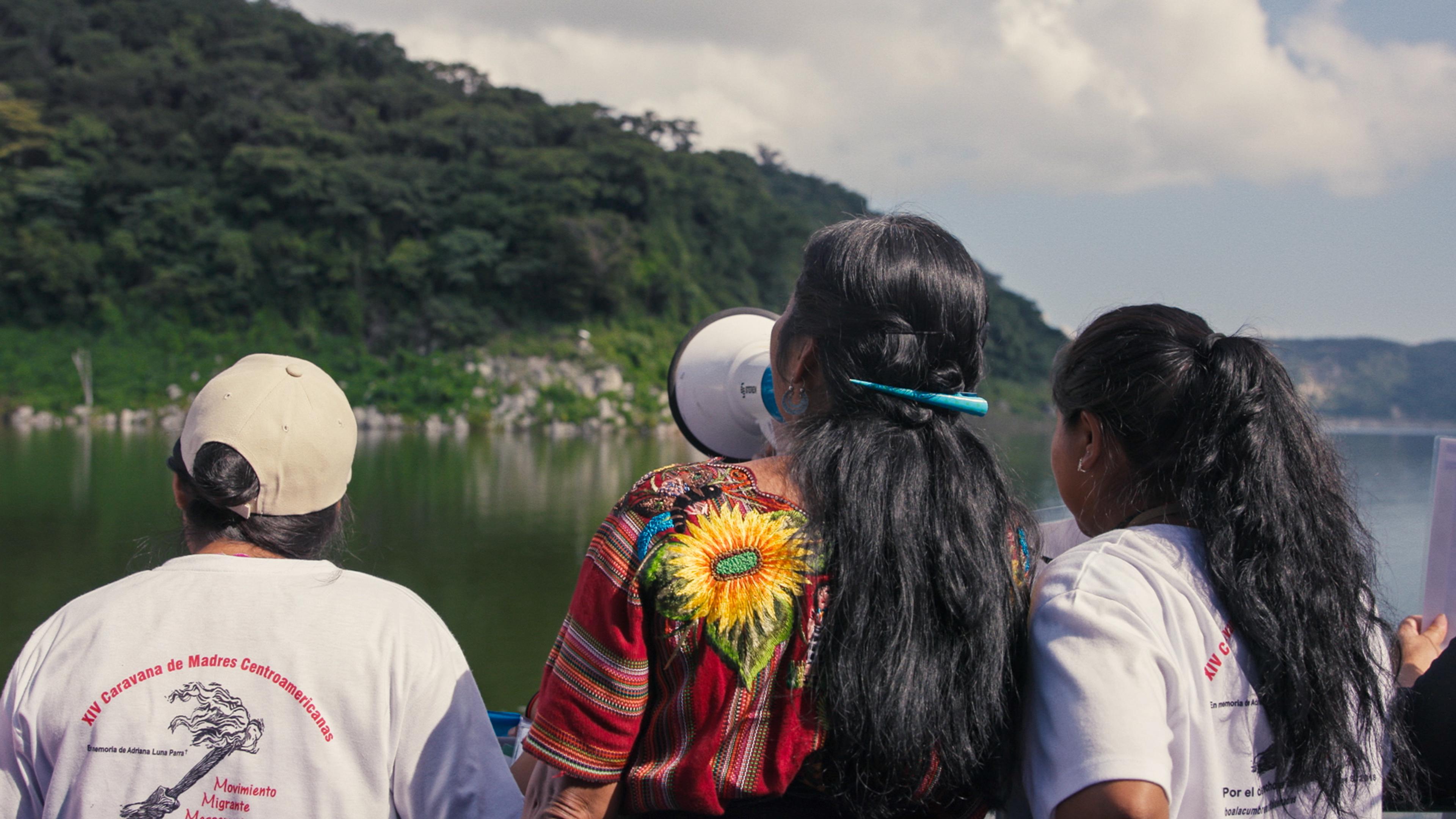 Photo of three women viewed from behind near a lake with trees. One holds a megaphone and wears a colourful embroidered shirt.