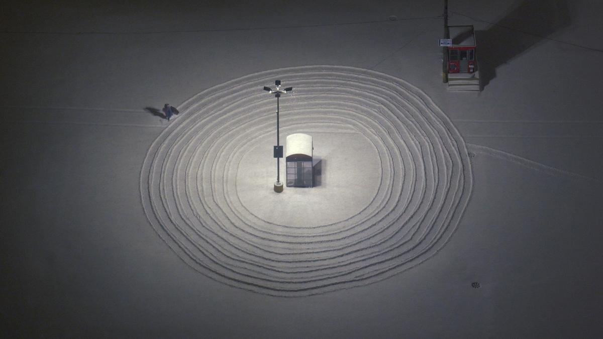 Aerial view of concentric snow tracks around a phone booth and street light. The artist walks in circles away from the center, dragging a suitcase behind her.