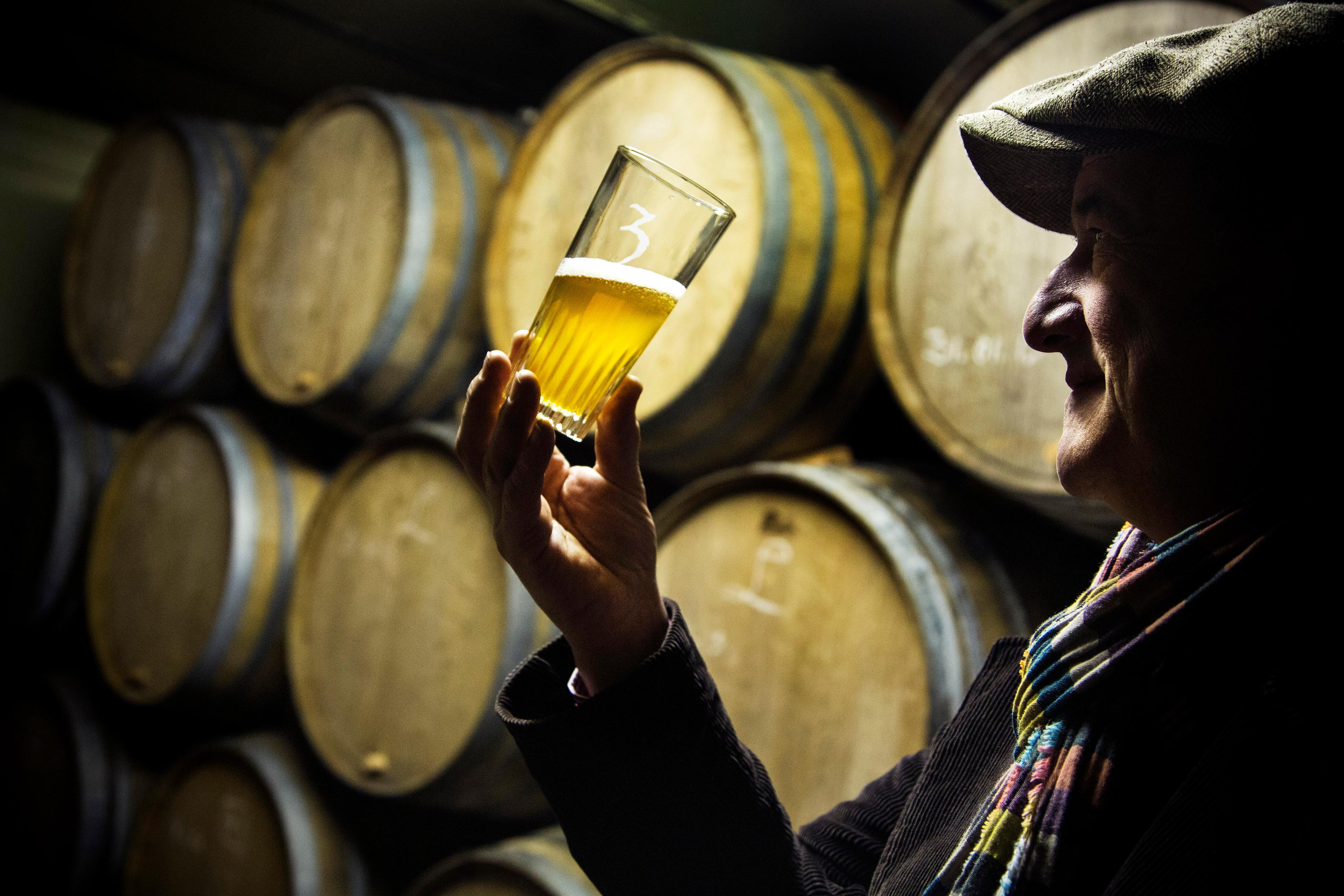 Photo of a man in a hat inspecting a beer glass in front of wooden barrels in a dimly lit room.