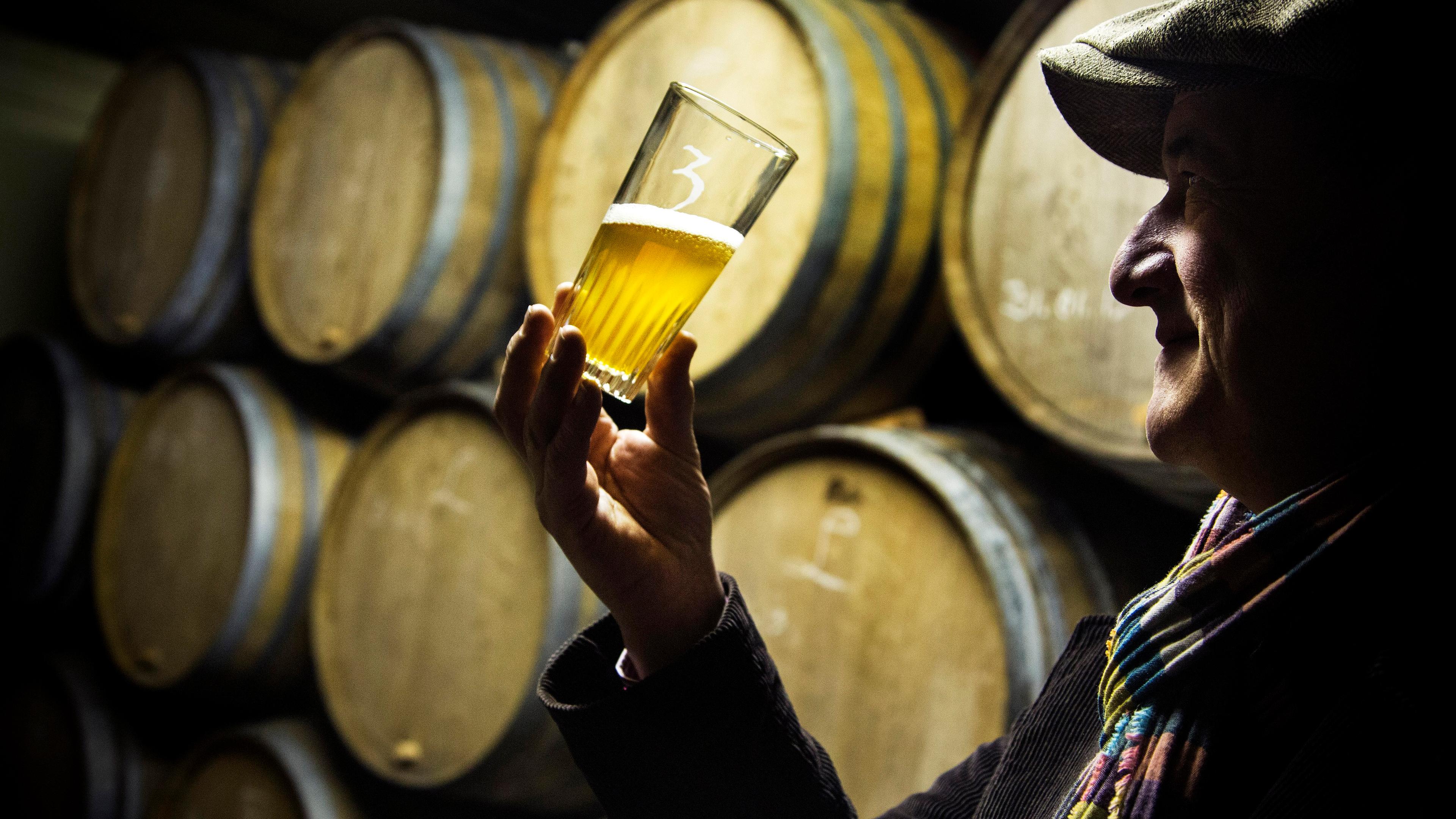 Photo of a man in a hat inspecting a beer glass in front of wooden barrels in a dimly lit room.