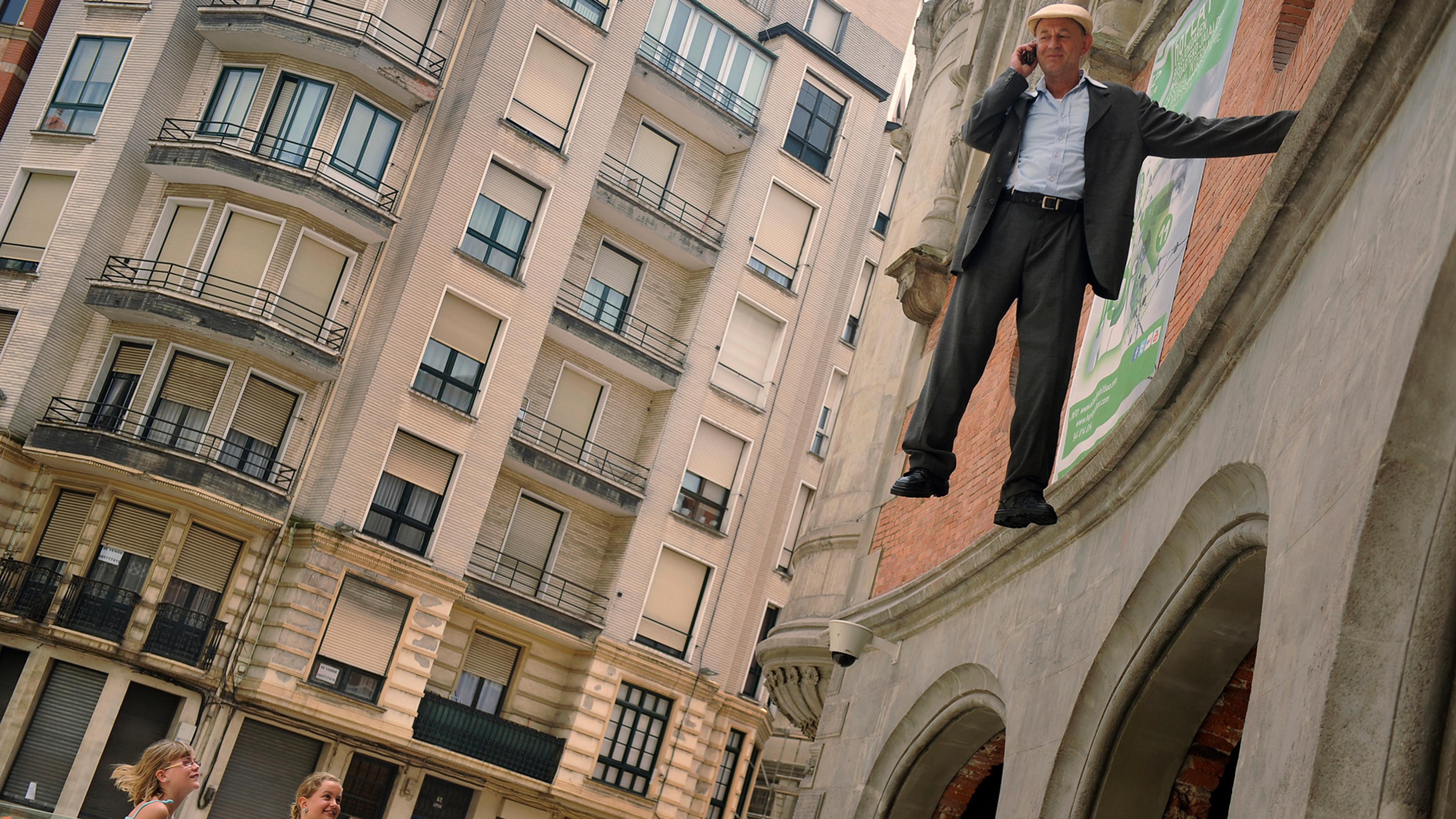 A man in a suit and hat floats above the heads of onlookers beside an office building, holding a mobile phone.
