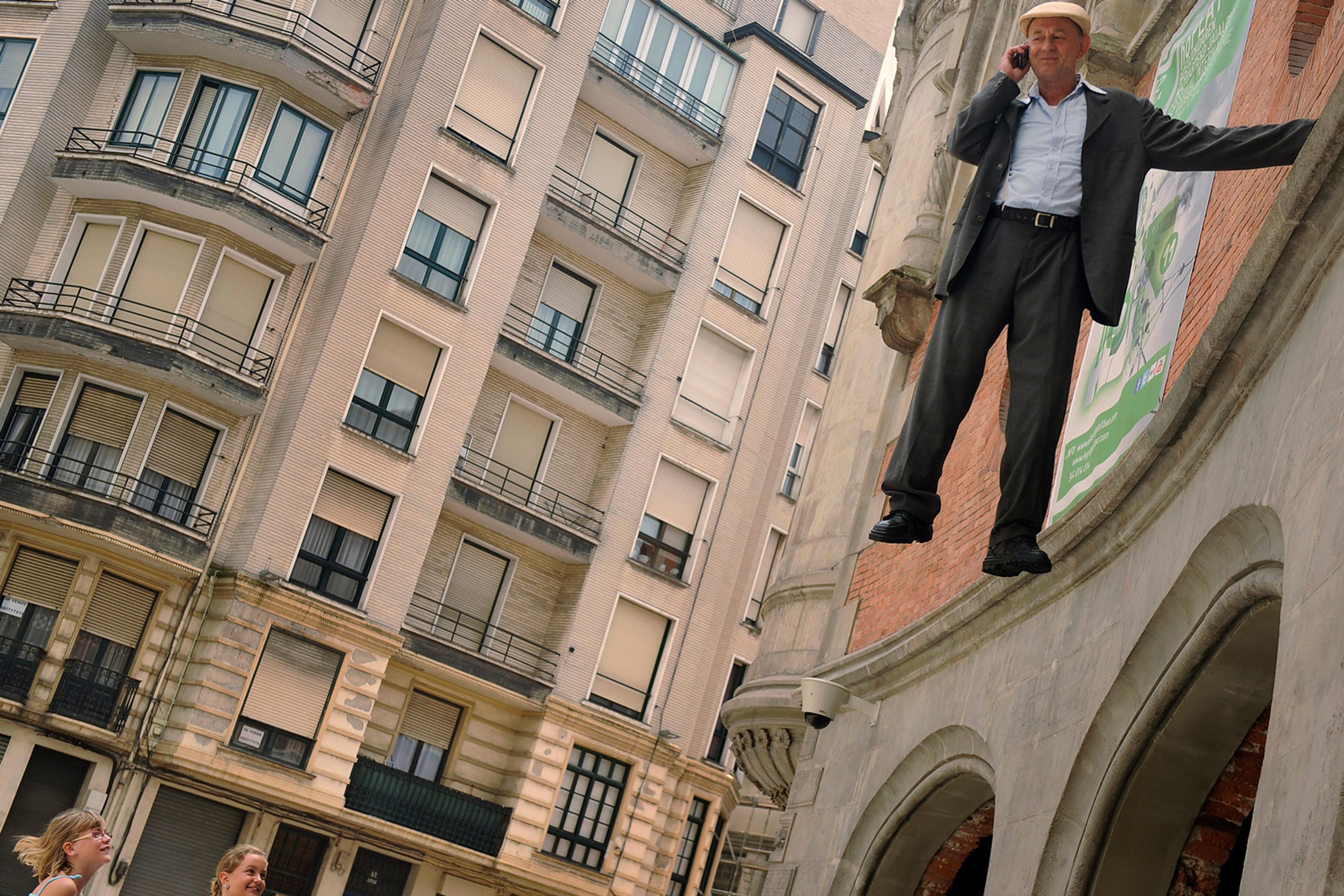 A man in a suit and hat floats above the heads of onlookers beside an office building, holding a mobile phone.