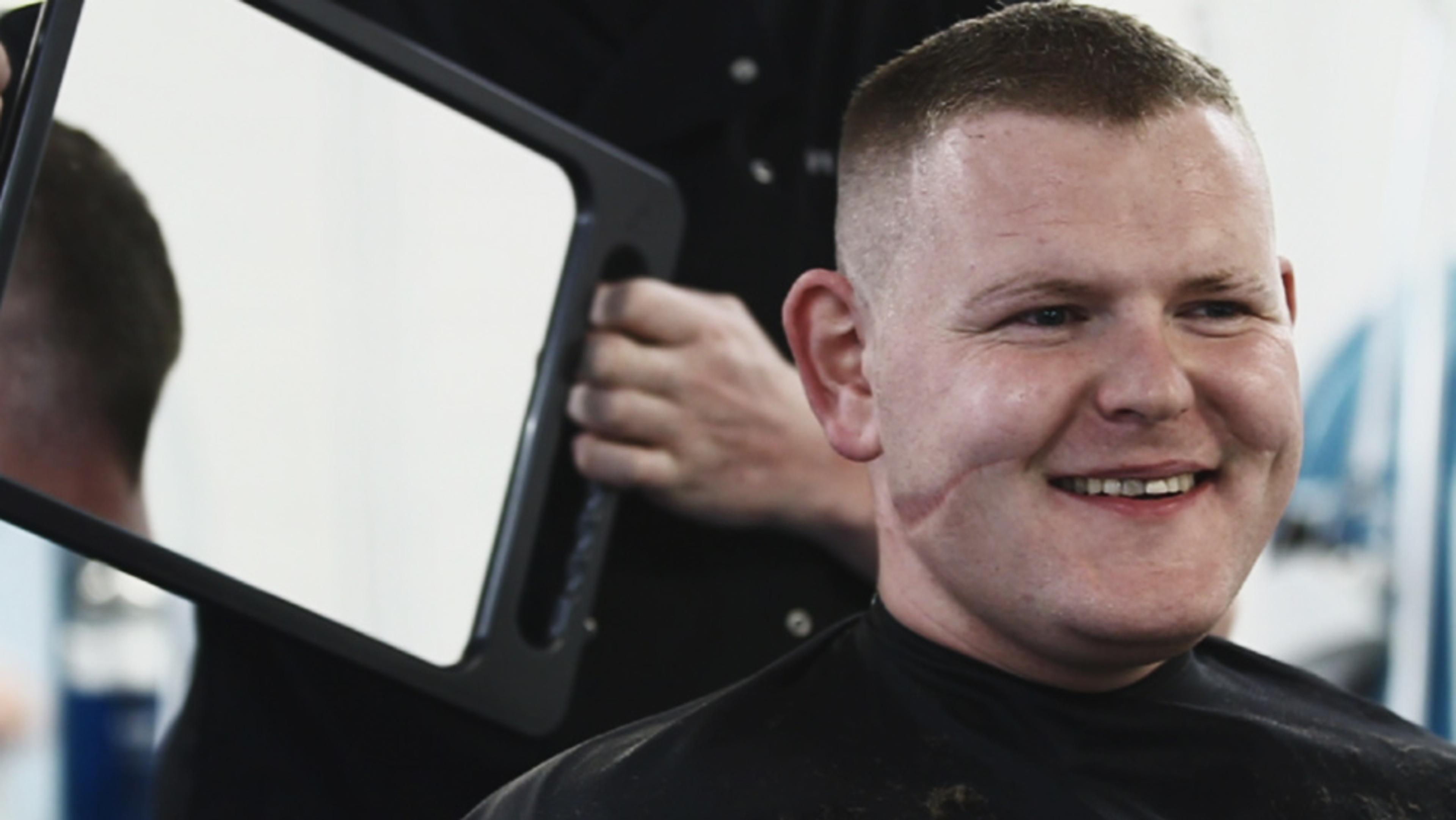 A smiling man with short hair in a barber’s chair while the barber holds up a mirror for him to see the haircut.