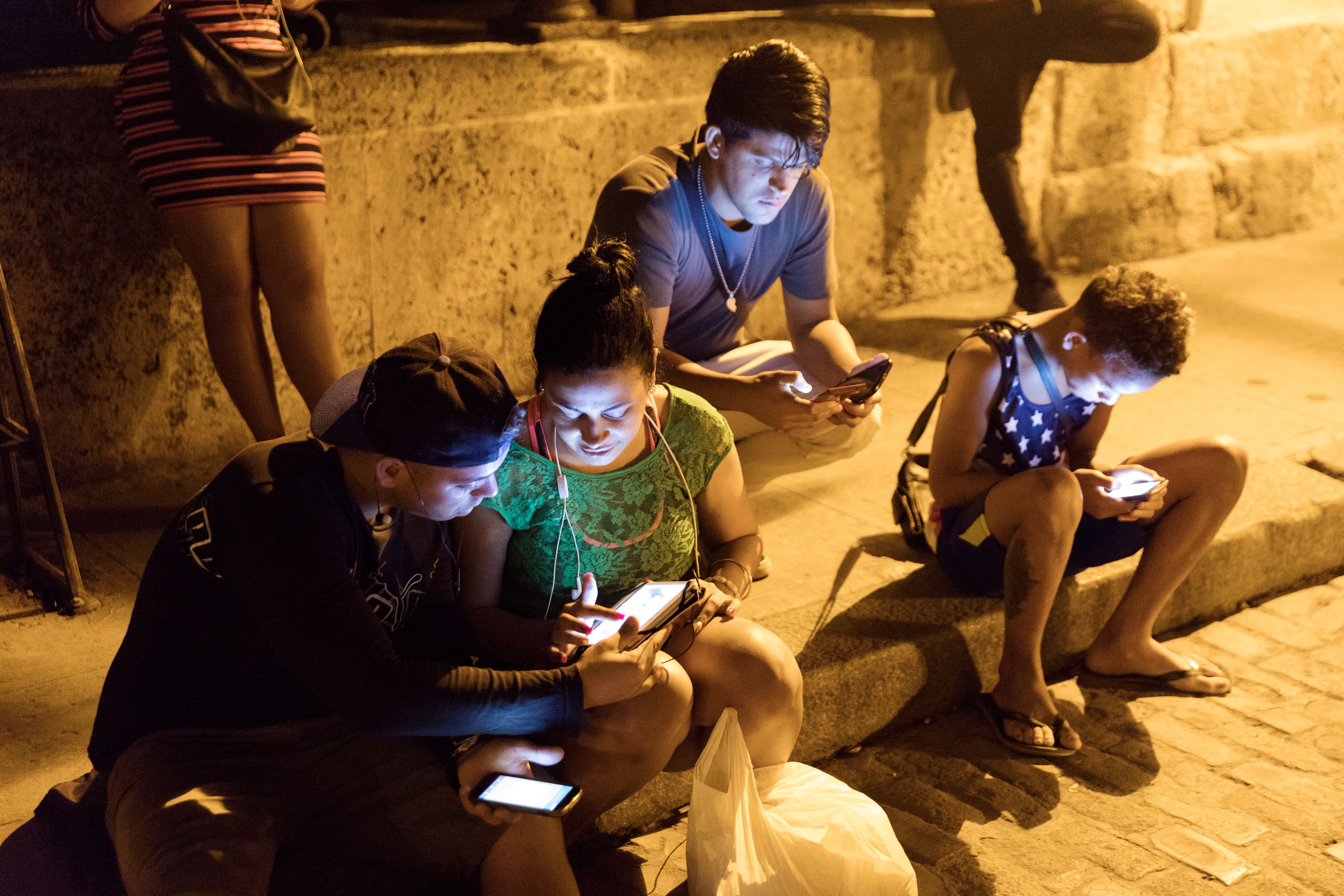Photo of people sitting on a street using smartphones at night, illuminated by screen light.