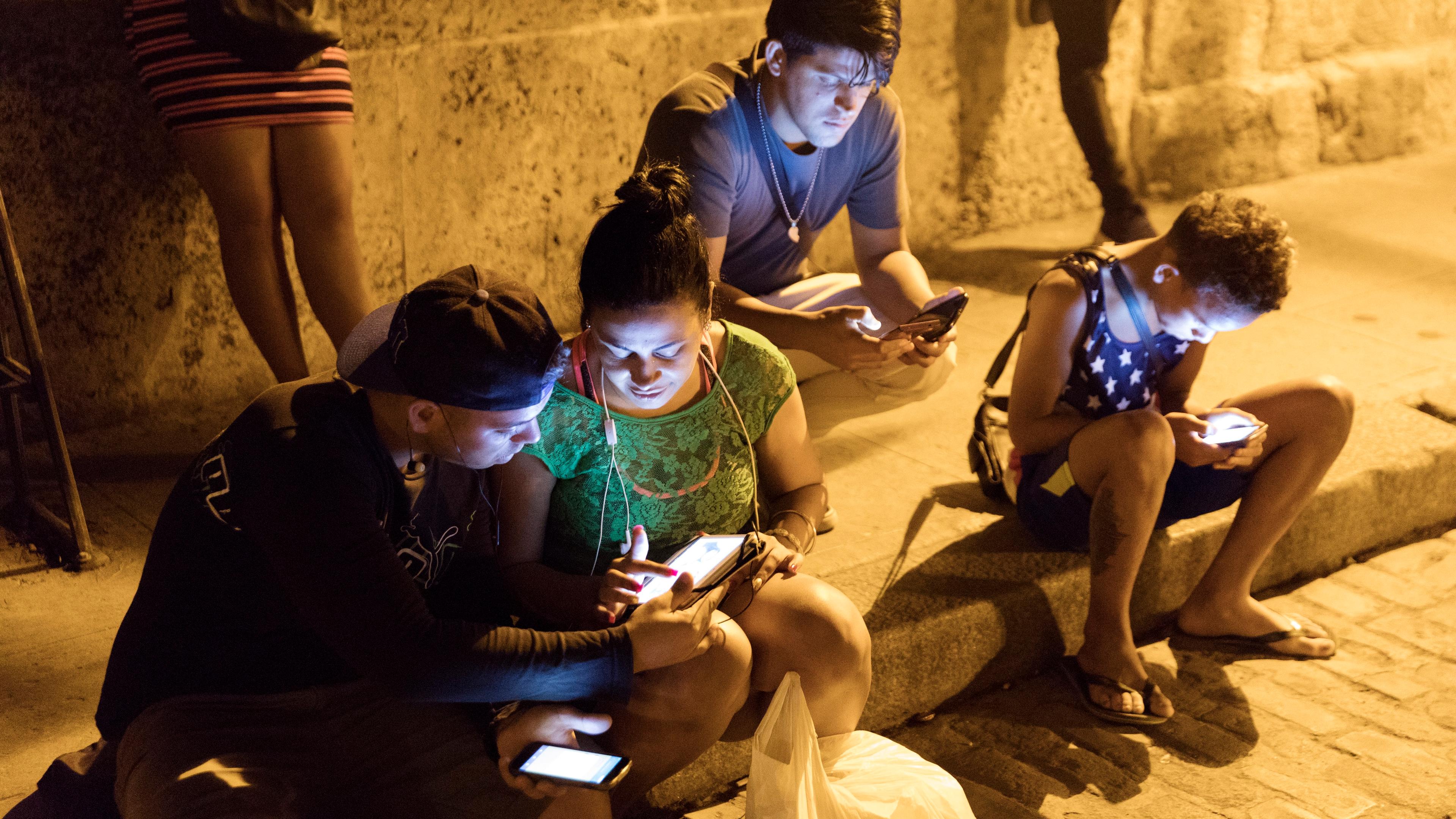 Photo of people sitting on a street using smartphones at night, illuminated by screen light.