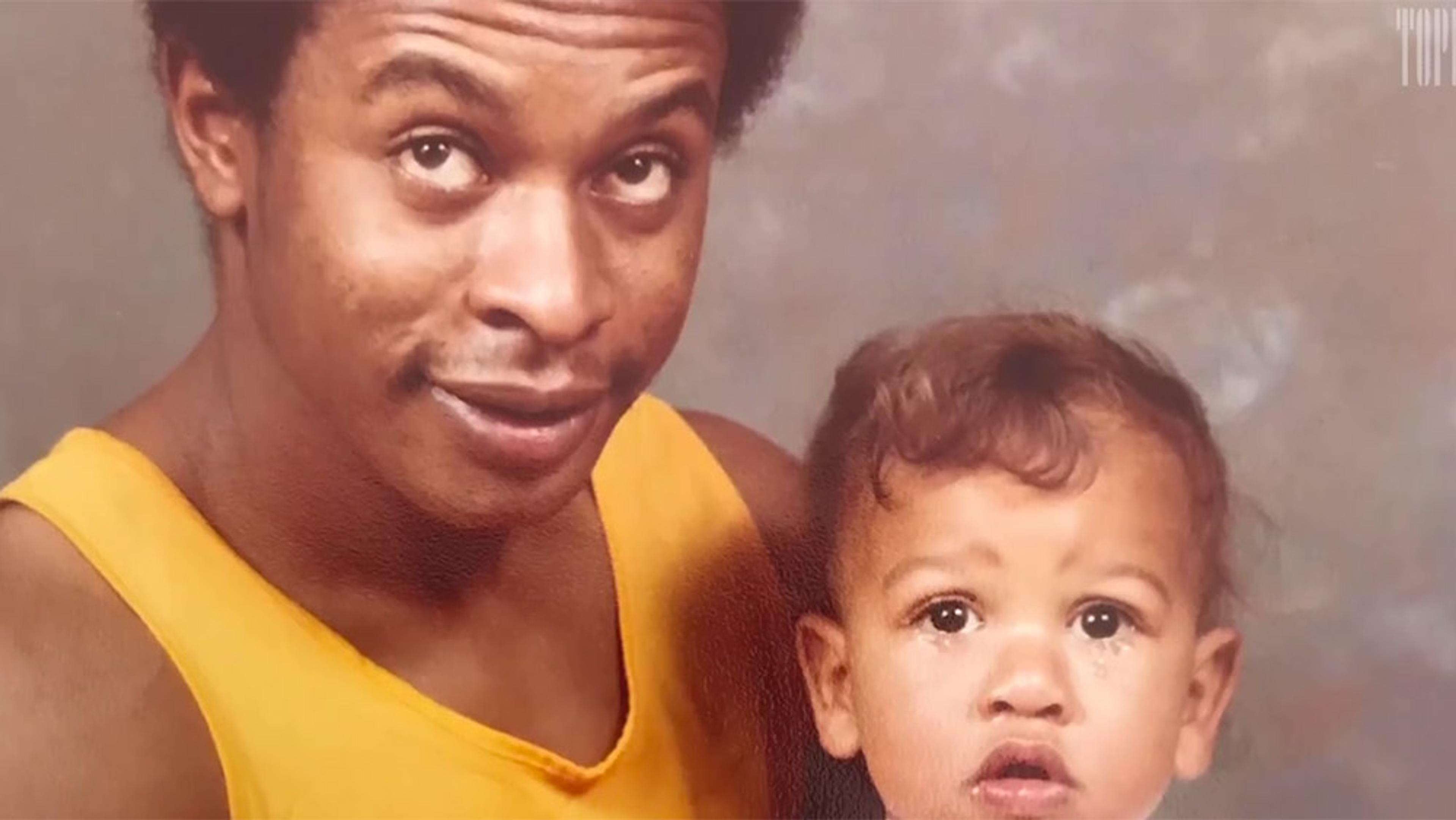 A Black man in a yellow tank top holding a child with a surprised expression against a neutral background.
